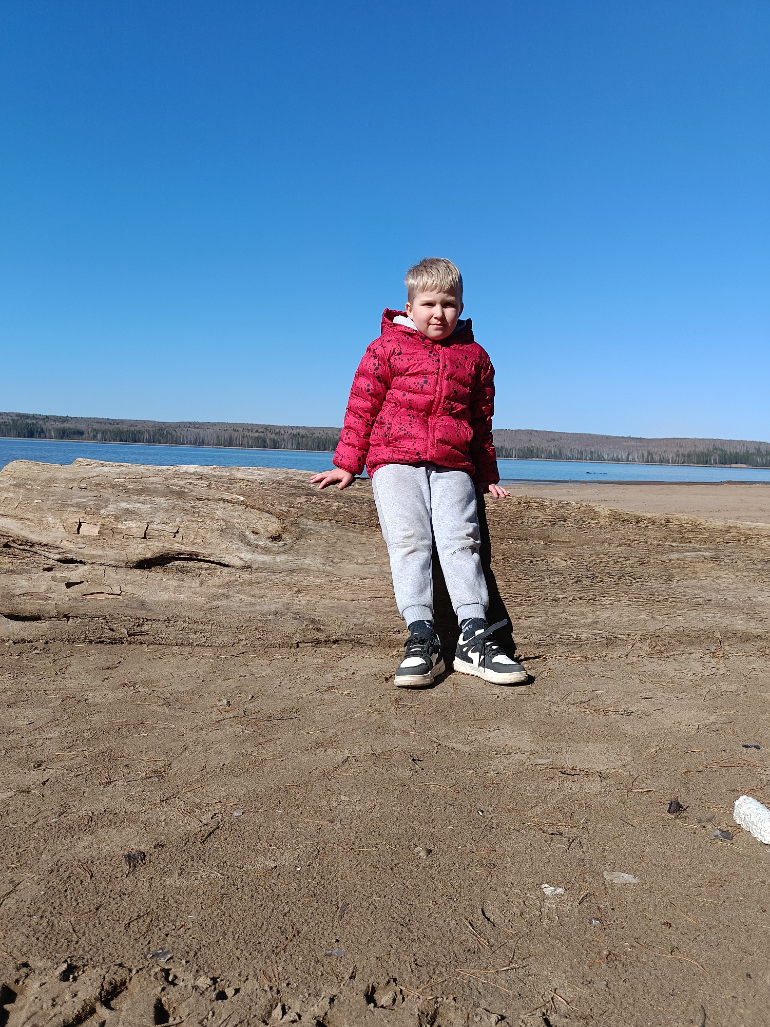 A full shot captures a young child with blond hair, wearing a red puffer jacket, gray sweatpants, and black and white sneakers. The child is positioned on a large, weathered log, possibly driftwood, in an outdoor environment. Behind the child, there is a body of water with a treeline visible in the distance, under a clear blue sky. The location appears to be a sandy beach or shoreline. The city and location are not evident in the image.