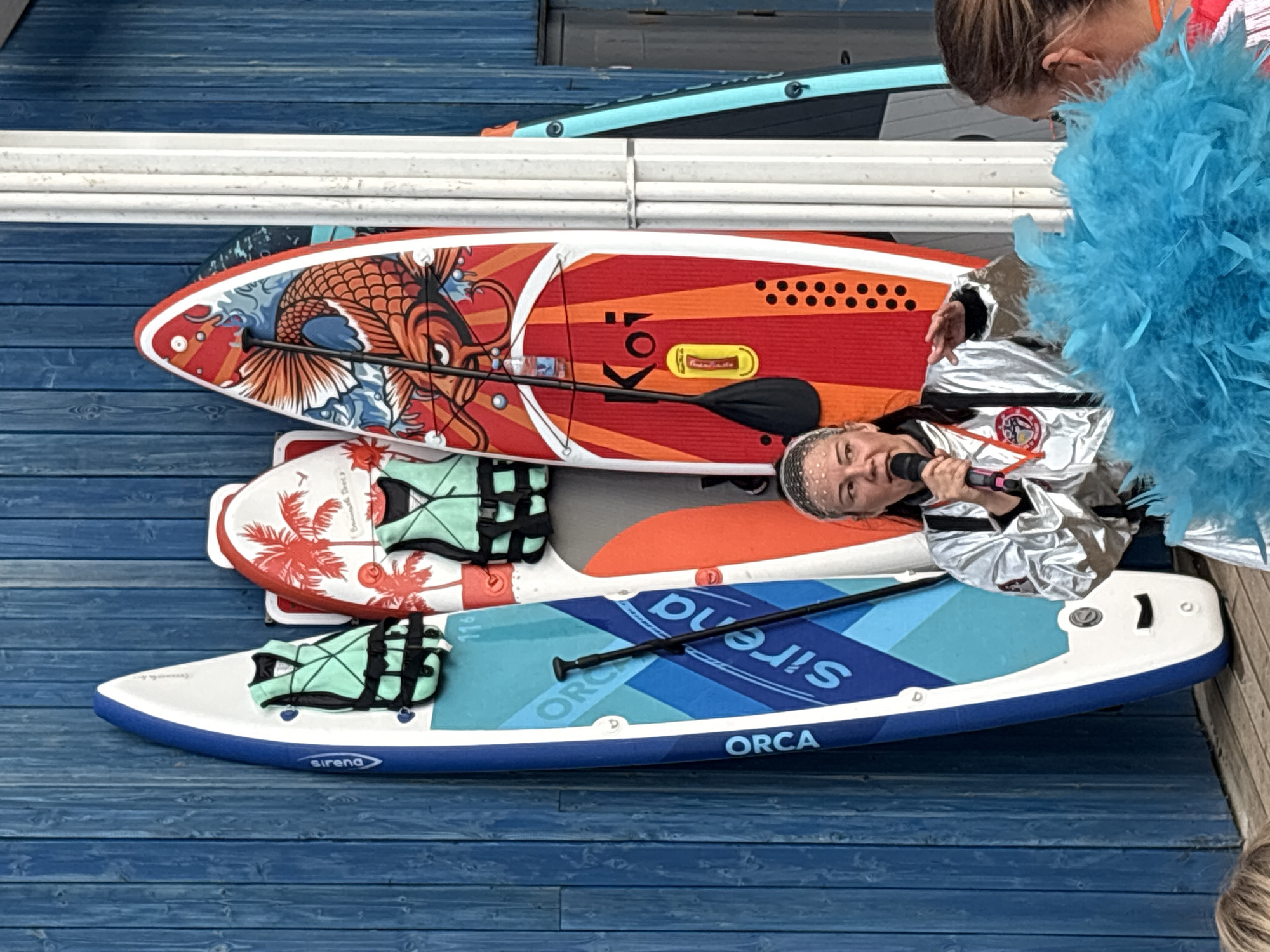 The image shows a close-up, overhead view of three stand-up paddleboards (SUPs) stacked on a blue wooden deck. The top SUP features a vibrant red and orange design with a stylized koi fish graphic. The middle SUP is white with a red palm tree design and two teal life vests. The bottom SUP is a lighter blue with darker blue stripes and "Sirena" and "ORCA" printed on it. A paddle is positioned across the top two boards.
A person in a shiny silver jacket is visible, holding a microphone and speaking into it. They are wearing a white head covering. Part of another person, with blonde hair, is visible at the top right of the frame, along with a large fluffy blue accessory that could be a boa or costume element. The lighting suggests it is daytime, and the overall mood seems to be one of an outdoor event or performance. The blue wooden deck and railing suggest a waterside location. The text "Sirena" and "ORCA" are visible on the bottom SUP.