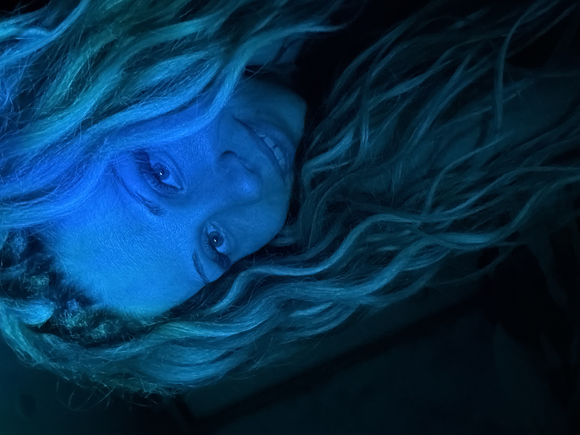 The image is a close-up portrait of a woman with blonde, wavy hair and braided sections near her forehead. She is looking towards the camera with a slight smile. The lighting is predominantly blue and teal, casting a dramatic effect on her face and hair. The background is dark and out of focus. There is no information in the image to suggest it is located in Rusanovo, Russia.