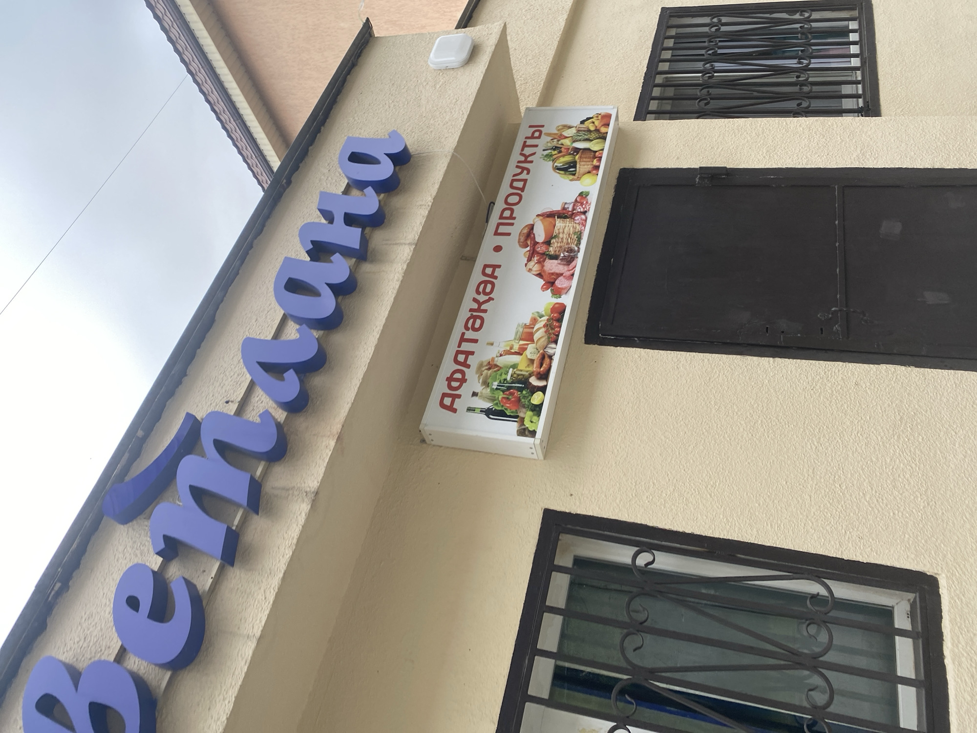 The image shows the exterior of a store. A large, blue sign with cursive lettering is mounted above the store's entrance. Below this, a rectangular sign displays images of food items and text in a foreign language. The store has a dark door and two windows with metal bars. The building's facade is painted a light yellow, and the roof is visible at the top of the image. The sky appears overcast.