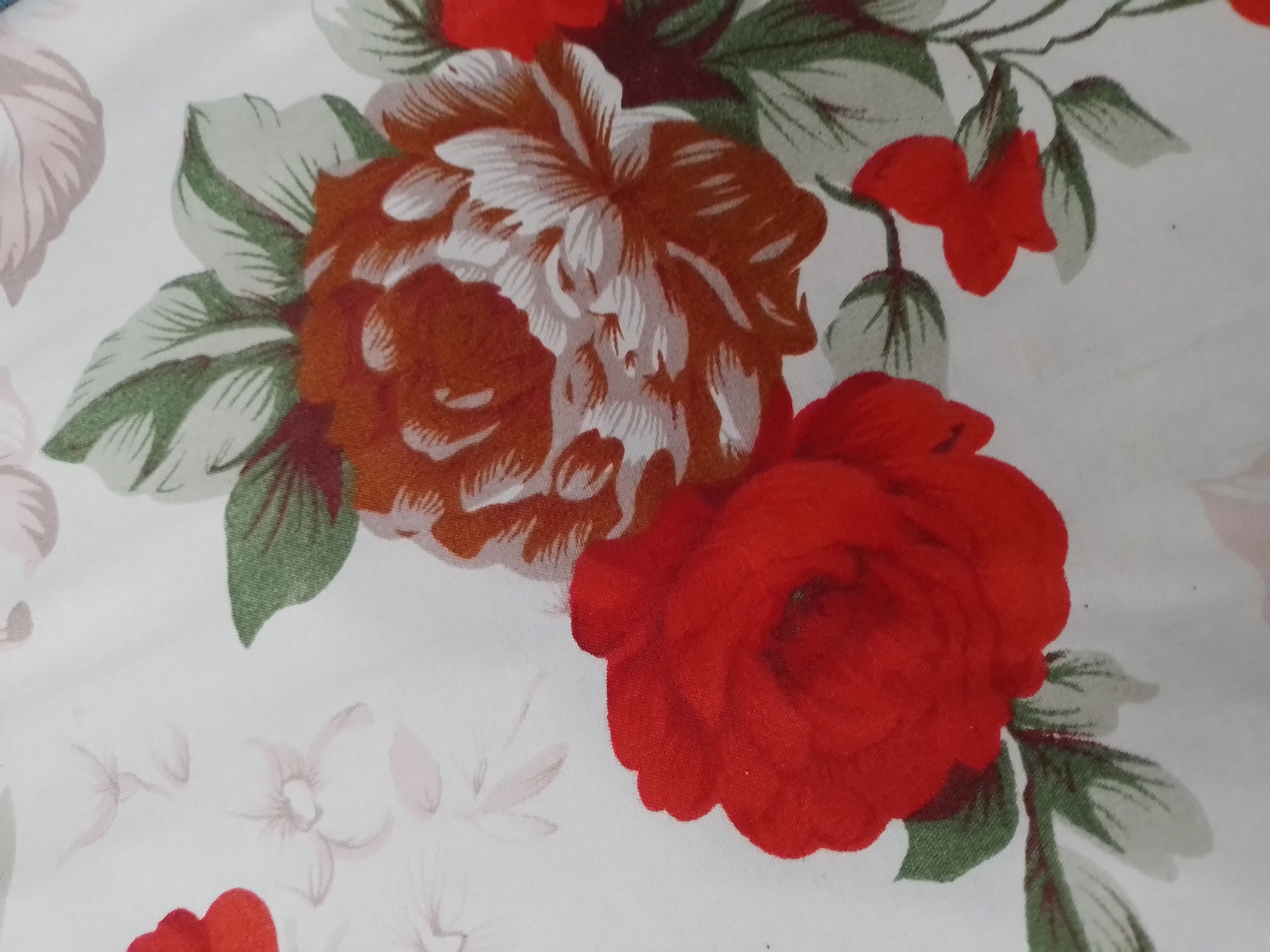 The image shows a close-up of a floral patterned fabric. The pattern features several roses, some in red and others in a brownish-orange and white combination, interspersed with green leaves and lighter, more abstract floral shapes in the background. The fabric appears to be a light color, possibly white or off-white, serving as the base for the print. There are no people or specific objects indicating a location within the city of Ngancar, Indonesia. The image is a detail shot of a textile design.