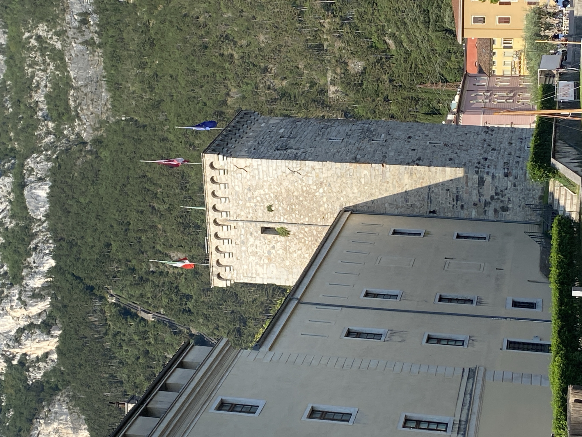 The image depicts a view of the Torre Apponale, a historic tower located in Riva del Garda, Italy. The tower, constructed of stone, stands prominently in the foreground, topped with crenellations and flying three flags: the Italian flag, a red and white checkered flag, and the European Union flag. To the left of the tower, a pale yellow building with multiple windows and architectural details extends into the frame. Behind these structures, a steep, forested mountainside rises, with a visible cable car track ascending its slope. In the distance, to the right of the tower, other buildings of varying colors are visible, suggesting an urban setting. The scene is illuminated by sunlight, casting shadows from the tower and buildings. A dense green hedge runs along the bottom of the image in front of the buildings, and a few indistinct figures and vehicles are present on a road in the far right background.