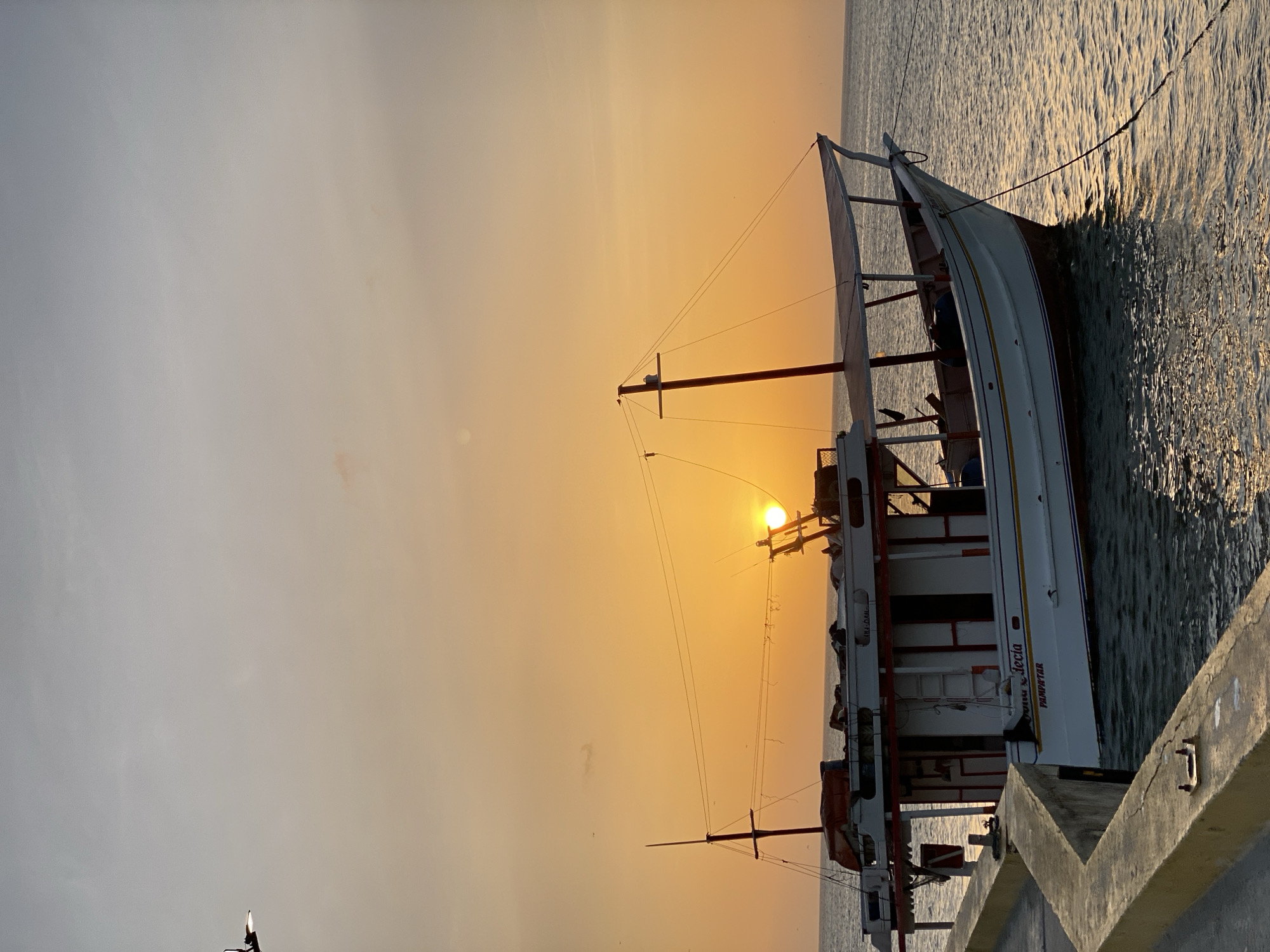 The image shows a white and red fishing boat docked at a concrete pier in Juan Griego, Venezuela. The sun is setting, casting an orange glow across the sky and water. The boat has a tall mast with rigging, and the name "Nuestra Decia" is visible on the side. The water is calm with light ripples. The pier is made of concrete and appears to be old.