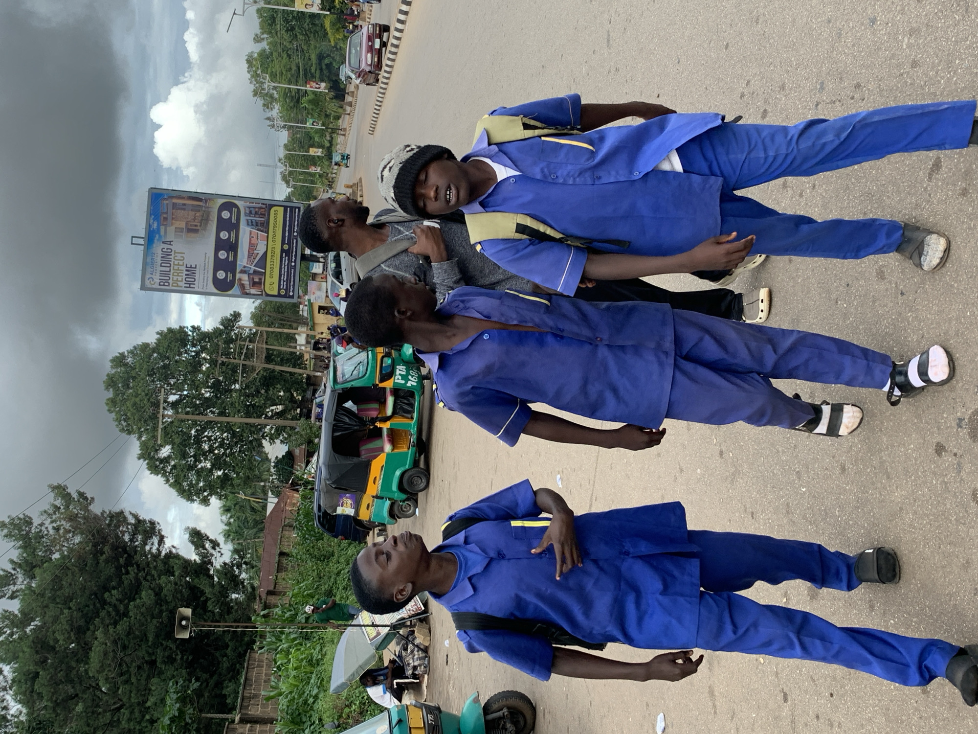 Three young boys in blue uniforms, two with backpacks, walk along a paved road in Barakin Lerawi, Nigeria. The boy on the far left wears a dark sandal and the other two are wearing sandals as well. The boy in the middle is wearing a beanie. In the background, a bright yellow and green auto-rickshaw is parked on the side of the road. A large billboard advertising apartments is visible further down the road. The sky is overcast with clouds, and trees line the side of the road. A few cars are driving on the road.