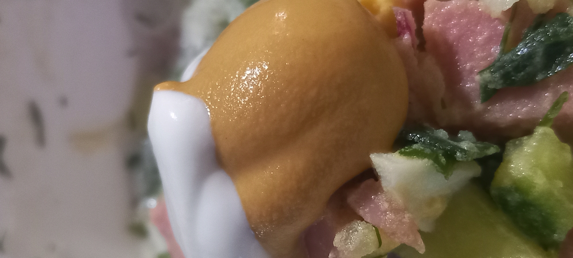 The image is a close-up, high-angle shot of a dish of food. A dollop of white sauce, possibly mayonnaise or sour cream, sits on top of a mound of golden-brown mustard. The mustard appears thick and textured, with some sheen from the lighting. Below the mustard is a salad composed of chopped pink ham, white pieces of what appear to be hard-boiled egg, and green elements that could be cucumber or herbs. The salad ingredients are mixed together and appear moist. The background is out of focus but suggests a tabletop or counter surface. There is no discernible information about the location within the city of Kryvyi Rih, Ukraine, in this image.