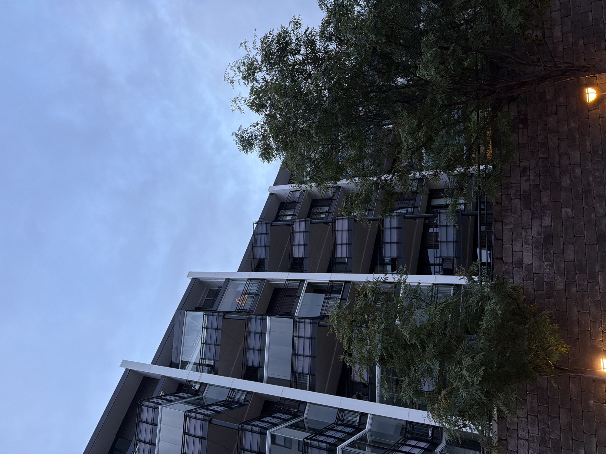 A modern apartment building with curved balconies and glass enclosures is viewed from a low angle, partially obscured by the foliage of two trees. A textured, dark brick wall runs along the foreground. The sky above is overcast and blue-gray. A string of lights, possibly a Christmas decoration, is visible on one of the balconies. Two streetlights with warm yellow illumination are positioned near the base of the wall. The building's architecture suggests it is located in a contemporary urban setting. Based on the visual style and typical modern architecture found in Norway, this building is likely situated in a city like Harstad, Norway, a city known for its modern infrastructure and scenic surroundings.