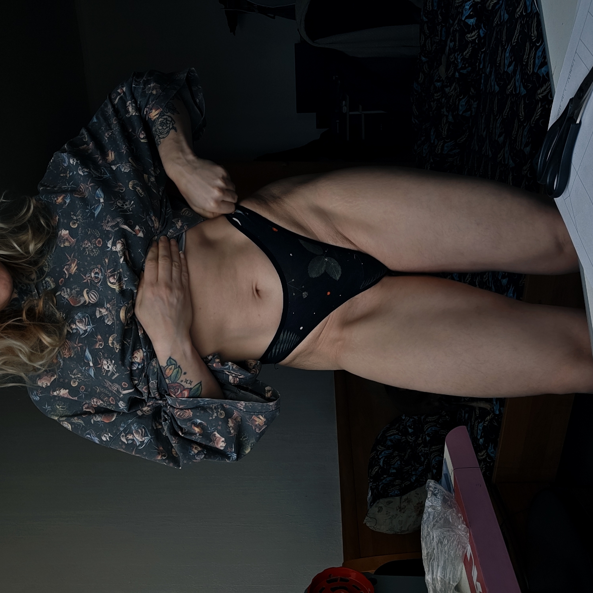 A medium shot depicts a woman with fair skin, wearing a patterned, loose-fitting top and black underwear with a subtle pattern of leaves or flowers. She is indoors, the lighting low, and is in the process of adjusting or holding the side of her underwear with her left hand. Her right hand rests on her stomach. Tattoos are visible on her right arm. A cluttered background is evident, with items including a patterned fabric or clothing item and a pair of scissors on top of a white surface. The location, Saint Petersburg, Russia, is not discernible from the visual information provided.