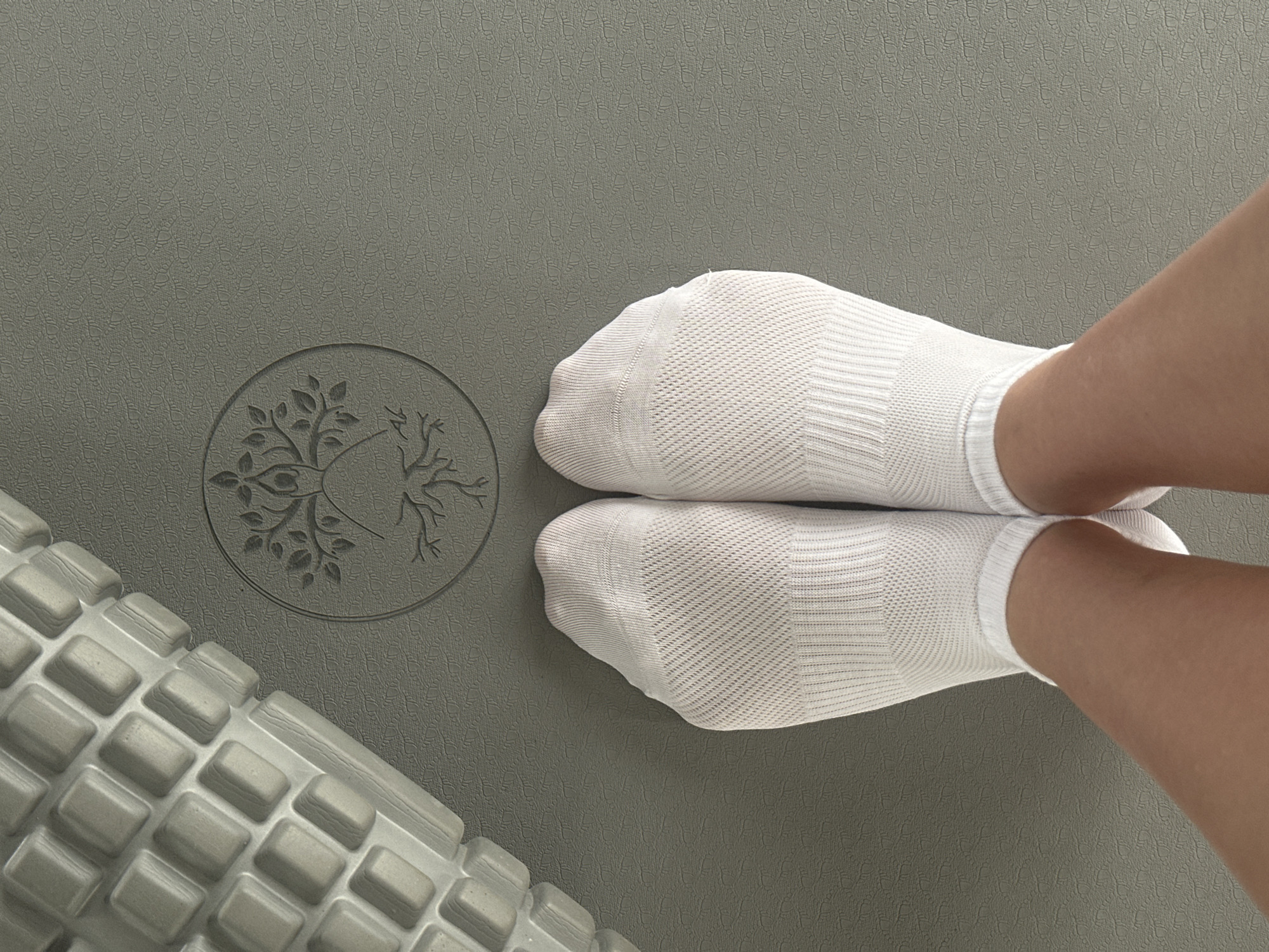 An overhead shot shows a person's feet, clad in white athletic socks, standing on a gray yoga mat. To the left of the feet is a textured foam roller, its cylindrical surface marked with a grid pattern of raised rectangles. Centered on the yoga mat, between the foam roller and the feet, is a circular emblem debossed into the mat's surface. The emblem depicts a stylized tree with outstretched branches, and at its base, a human-like figure with arms raised upwards, its form integrated into the tree's roots and trunk. The mat itself has a subtle, repeating texture of small, raised circular patterns. The scene is lit from above, creating soft shadows. There is no discernible connection to the city of Chaikovsky, Russia, within the image itself.