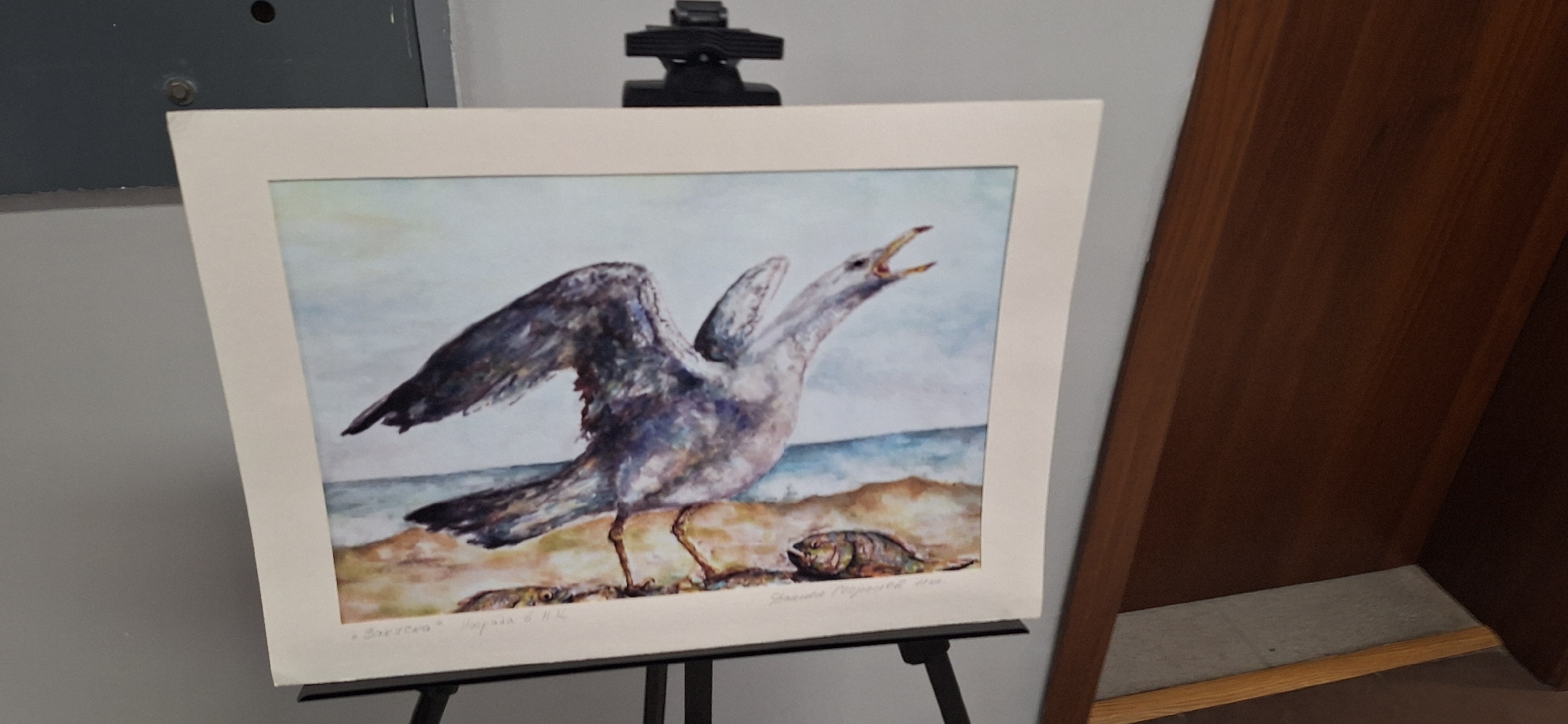 The image shows a framed watercolor painting of a seagull standing on a beach. The bird has its wings partially spread and its beak open as if calling out. There is a dead fish lying on the sand near its feet. The background of the painting depicts a beach and the sea. The painting is on display on a black easel in what appears to be an indoor setting. The information "Зовеска" Бургас 6 14г, Stanislava Neykova 1986" is visible on the lower edge of the painting. This suggests the painting could be found in Burgas, Bulgaria. A portion of a wooden door is visible on the right side of the image.