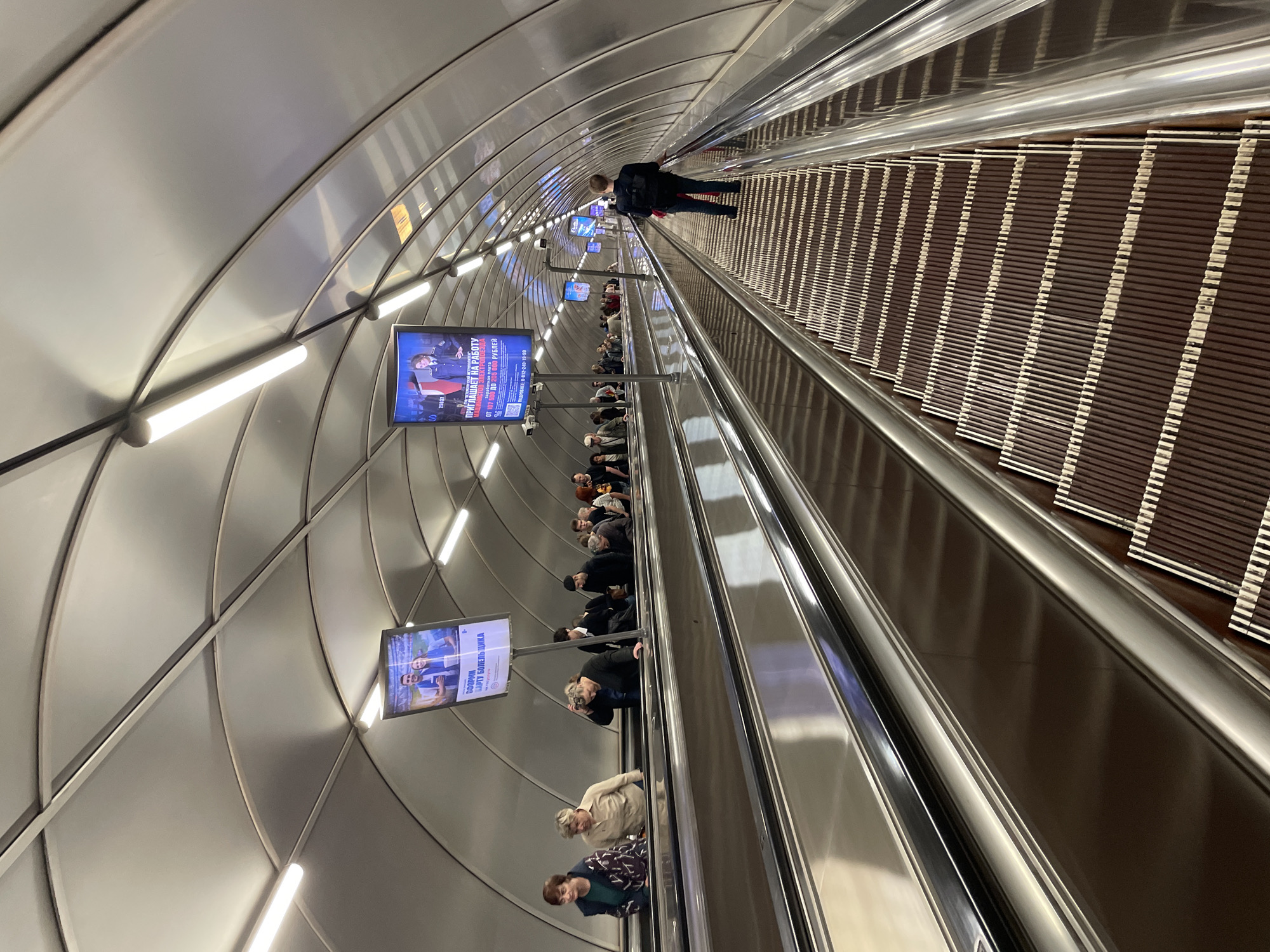 The image shows an escalator in a subway station in Saint Petersburg, Russia. The escalator is moving upwards, and several people are riding it. The station has a modern, curved ceiling with integrated lighting. On the left side of the escalator, there are two digital advertisements displaying images and text. The advertisement on the left shows a couple and text in Russian that appears to be about fan support. The advertisement on the right shows a person in a uniform and text in Russian that seems to be a job advertisement. A man in a dark jacket and backpack is walking up the escalator on the right side, facing away from the camera. The escalator steps are made of dark material with reflective stripes. The overall environment suggests a busy public transportation hub.