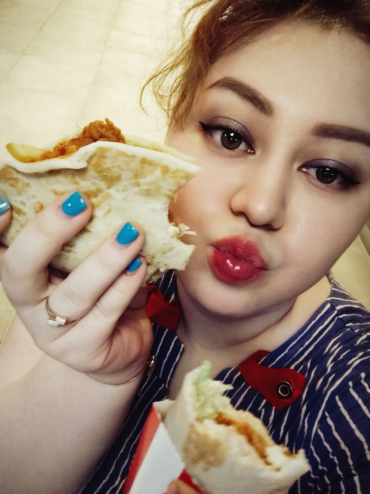 A close-up selfie captures a young woman with reddish-brown hair and striking eye makeup, puckering her lips as she holds a partially eaten wrap. She wears a blue and white striped shirt adorned with a red ribbon detail on the collar. Her right hand, adorned with bright blue nail polish and a diamond ring, is holding the wrap to her mouth. Another partially eaten wrap is visible at the bottom of the frame. The background appears to be an indoor tiled floor. The image was taken in Fergana City, Uzbekistan.