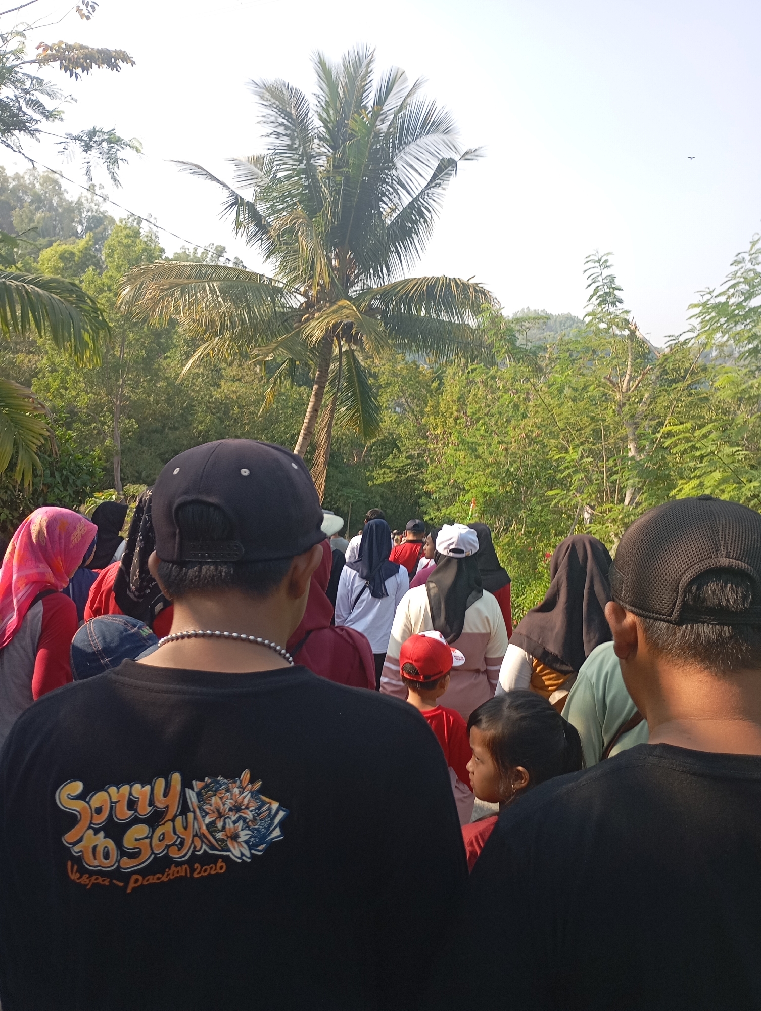 In Ketepung, Indonesia, a group of people, mostly wearing casual clothing and some with headscarves, are walking outdoors on a sunny day. The foreground is dominated by the backs of two individuals wearing black t-shirts. The person on the left has a black baseball cap and a beaded necklace, and their t-shirt displays a graphic with the words "Sorry to Say!" and "Vespa - pacitan 2026". The person on the right is also wearing a black t-shirt and a black baseball cap, seen from the back. In the middle ground, a child with dark hair tied in a ponytail, wearing a red shirt and a red baseball cap, is visible, looking to the left. Other individuals in the group are wearing a variety of clothing, including headscarves in pink, black, and white, and shirts in red, white, and cream. The environment is lush and green, with numerous trees, including a prominent palm tree, and a hint of a hillside in the background. The sky is bright and clear.