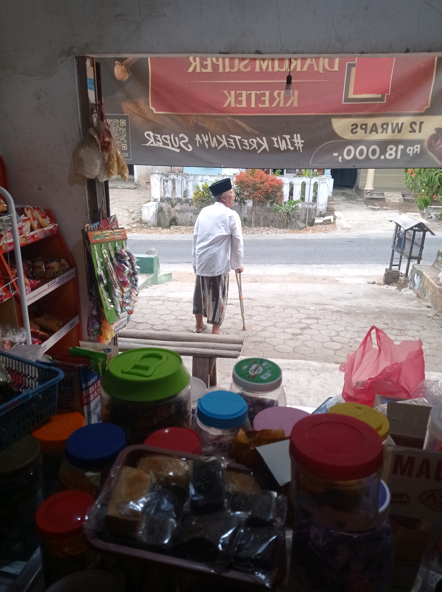 The image captures a scene from the doorway of a small shop in Purwosekar, Indonesia. In the foreground, an assortment of jars and containers with colorful lids are visible, suggesting the shop sells various goods, possibly food items or ingredients. A basket with blue plastic handles is also present.
Through the open doorway, a street scene unfolds. A lone elderly man, dressed in a white shirt and traditional patterned pants, stands with the aid of a walking stick. He faces away from the viewer, looking towards the street and a sidewalk paved with hexagonal tiles. Beyond the sidewalk, a road is visible, with a few buildings and some vegetation in the background. A red plastic bag rests on the sidewalk to the right.
Above the doorway, a banner displays text in Indonesian. The upper portion of the banner reads "DIJUAL SUPER KRETEK," and below that, "#INI KRETEK". Another section of the banner shows a price of "RP 18.000,0". The shop appears to be a local convenience store or a small general goods retailer situated in a residential or mixed-use area.