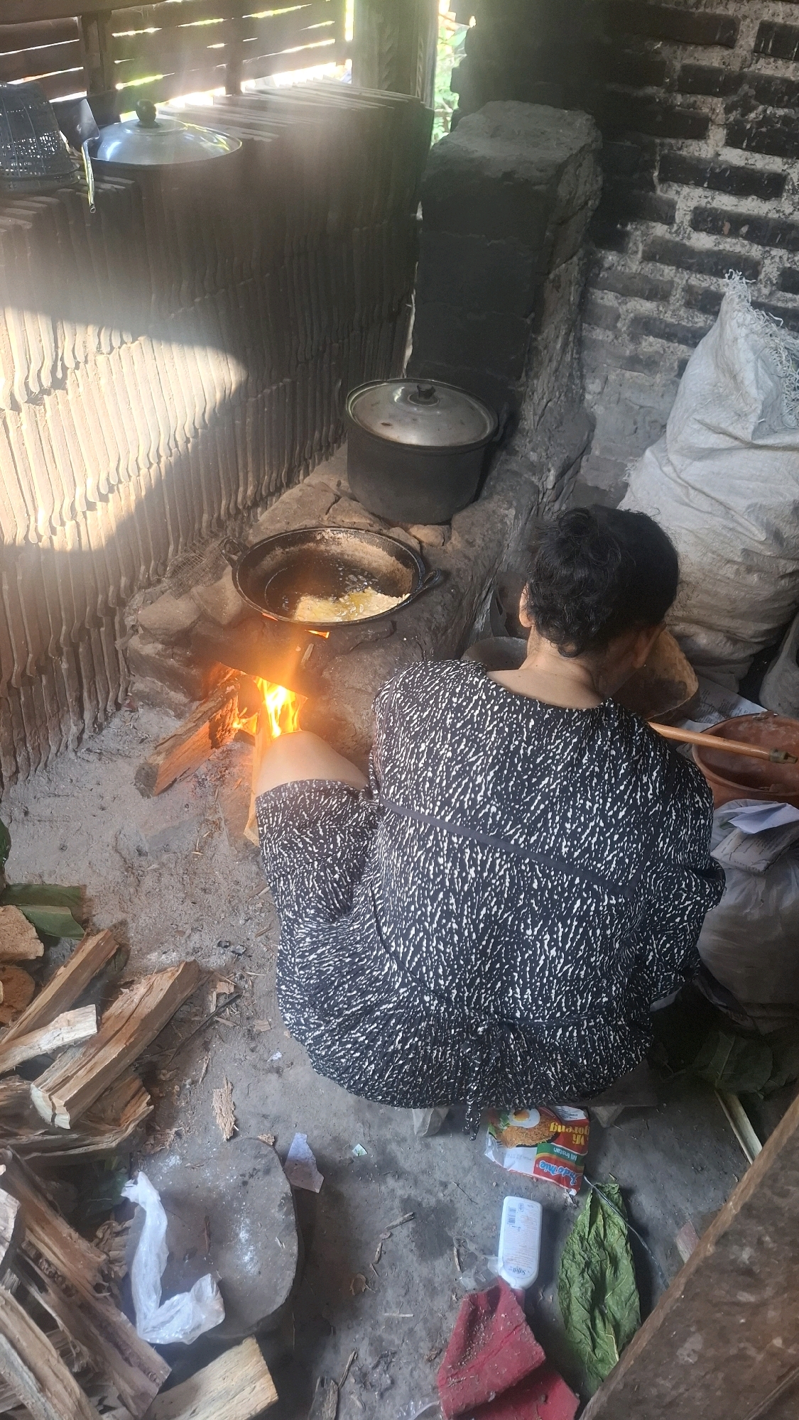 In Semanding, Indonesia, a person is crouched on the ground, facing away from the camera, and tending to a wood-fired stove. The person is wearing a black and white patterned dress. The stove is active with flames visible, heating a dark-colored pan filled with oil and food, possibly frying. Around the stove, there are several large sacks, firewood, and some banana leaves scattered on the dirt floor. In the background, there is a brick wall and what appears to be a traditional tiled roof structure. The overall scene suggests a rustic, outdoor cooking environment.