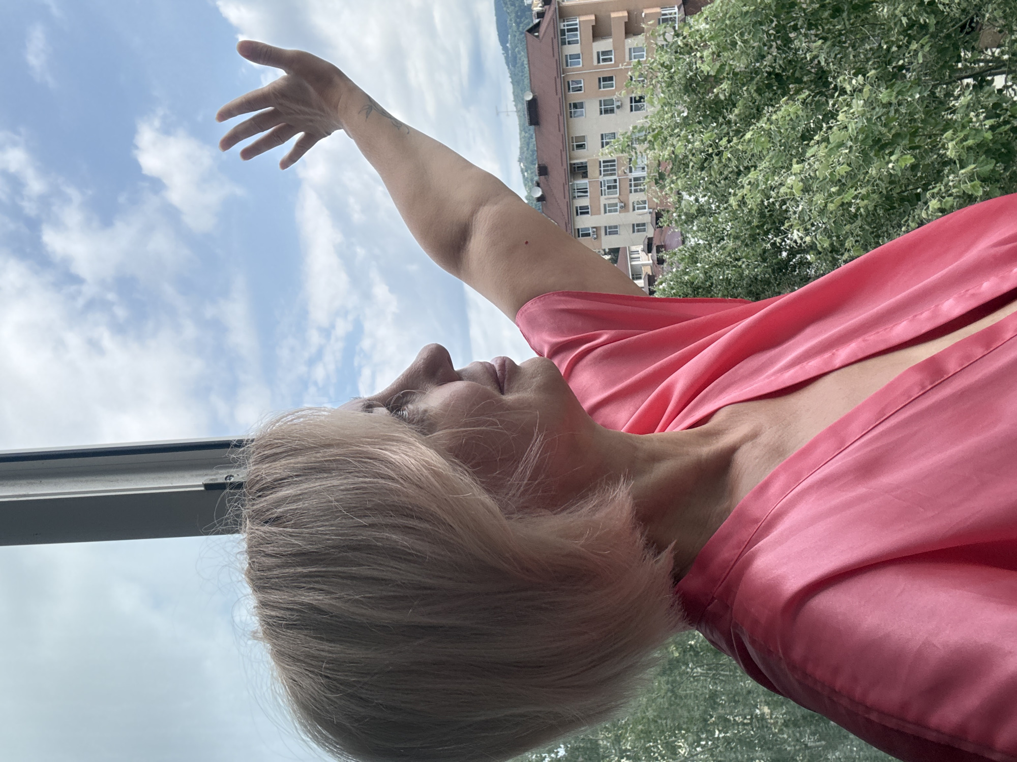 This is a slightly tilted, close-up, waist-up shot of a woman standing on a balcony in Sochi, Russia. She has short, blonde hair and is wearing a pink satin robe. Her left arm is extended upwards, with her hand open, reaching towards the sky. Her face is turned upwards and to the left, with her eyes closed and a serene expression. The background shows a bright, partly cloudy sky, lush green trees, and a building with balconies. The time of day appears to be daytime, likely mid-morning or afternoon, given the bright natural light. The overall mood is peaceful and contemplative, with the woman seemingly enjoying the fresh air and the view. There is no visible text in the image.