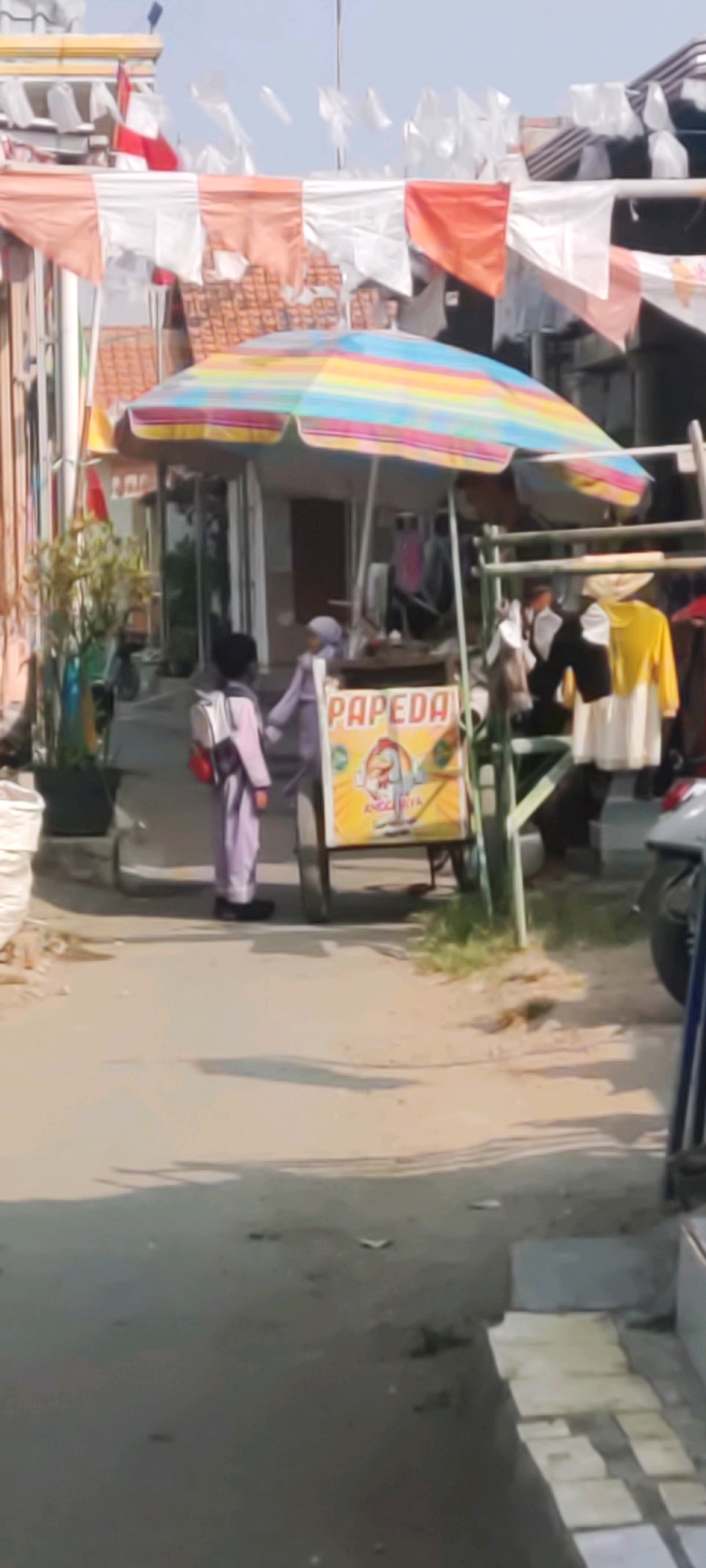 In the city of Gebang, Indonesia, a street scene unfolds under a striped umbrella. Two individuals are present. One, likely a child, is wearing a light purple outfit and a white backpack with a red strap. They stand facing another person, who is dressed in a light purple hijab and top, and is partially obscured by a cart. The cart has a prominent yellow sign with red lettering that reads "PAPEDA". The background shows buildings with flags and banners, suggesting a festive atmosphere. In front of the cart, clothing items are displayed on a rack, including a yellow top over a white garment. A motorcycle is parked to the right, and the ground is paved with a mixture of asphalt and concrete.