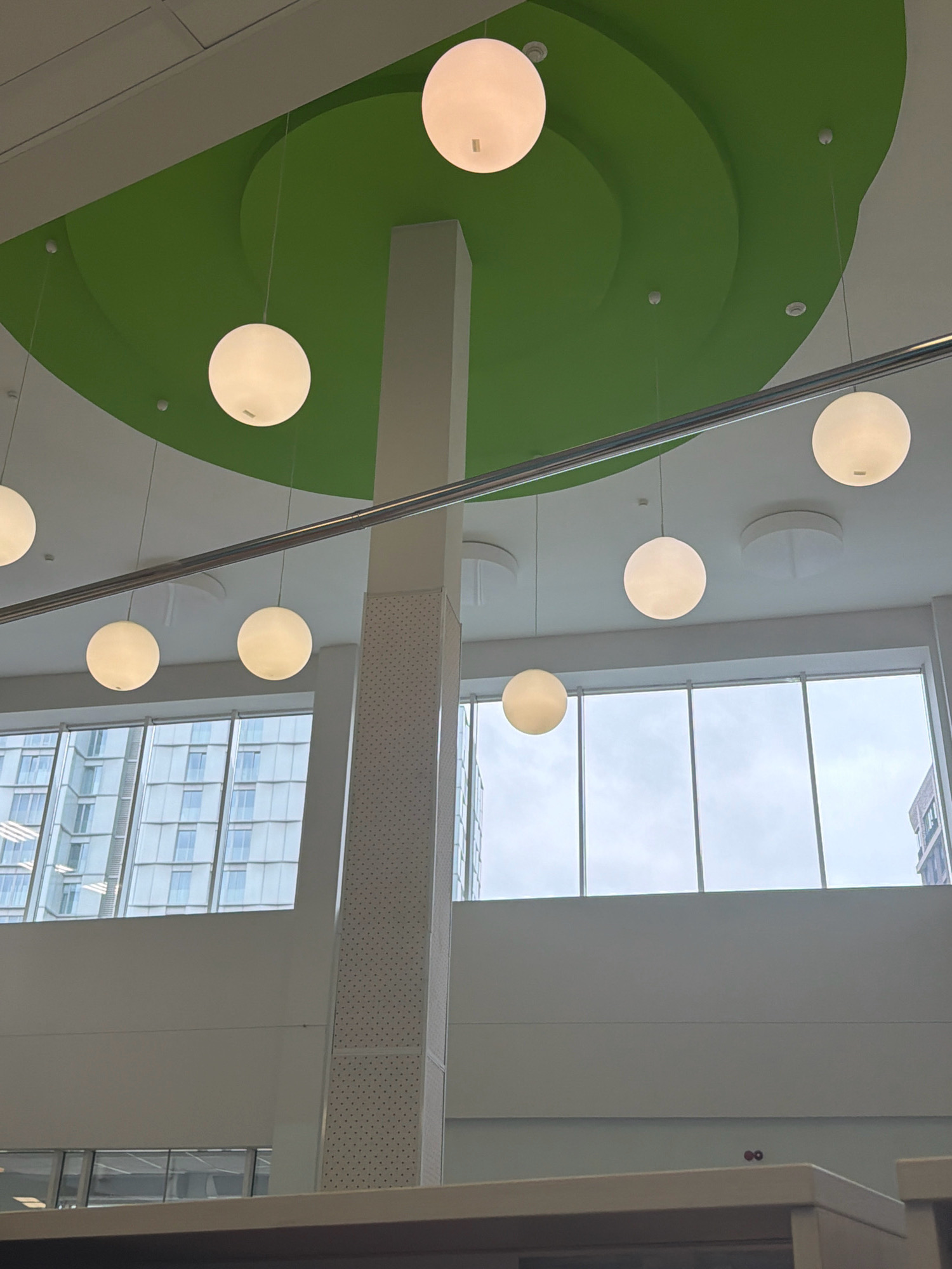 The image shows a modern interior space with a high ceiling and large windows overlooking an urban cityscape. The ceiling features a vibrant green, curved architectural element with recessed lighting and suspended spherical white pendant lights. A tall, rectangular column with a patterned surface divides the space. Through the expansive windows, several tall buildings with modern architecture and glass facades are visible. The overall impression is of a contemporary, well-lit environment, possibly a library, office, or public building. The specific location in Moscow, Russia cannot be determined from the visual information provided.