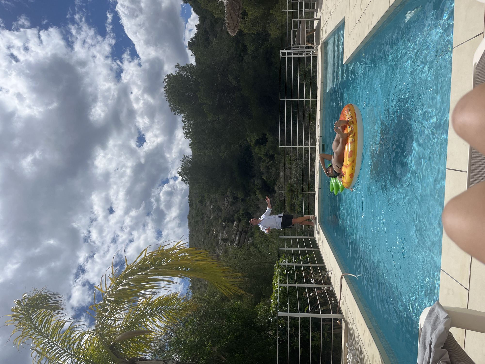The image depicts a swimming pool scene in El Poble Nou de Benitatxell, Spain, under a partly cloudy sky. A woman floats in the blue water on an inflatable ring. A man stands poolside, gesturing toward her. White fencing separates the pool area from a green hillside and vegetation. A palm tree is in the foreground.