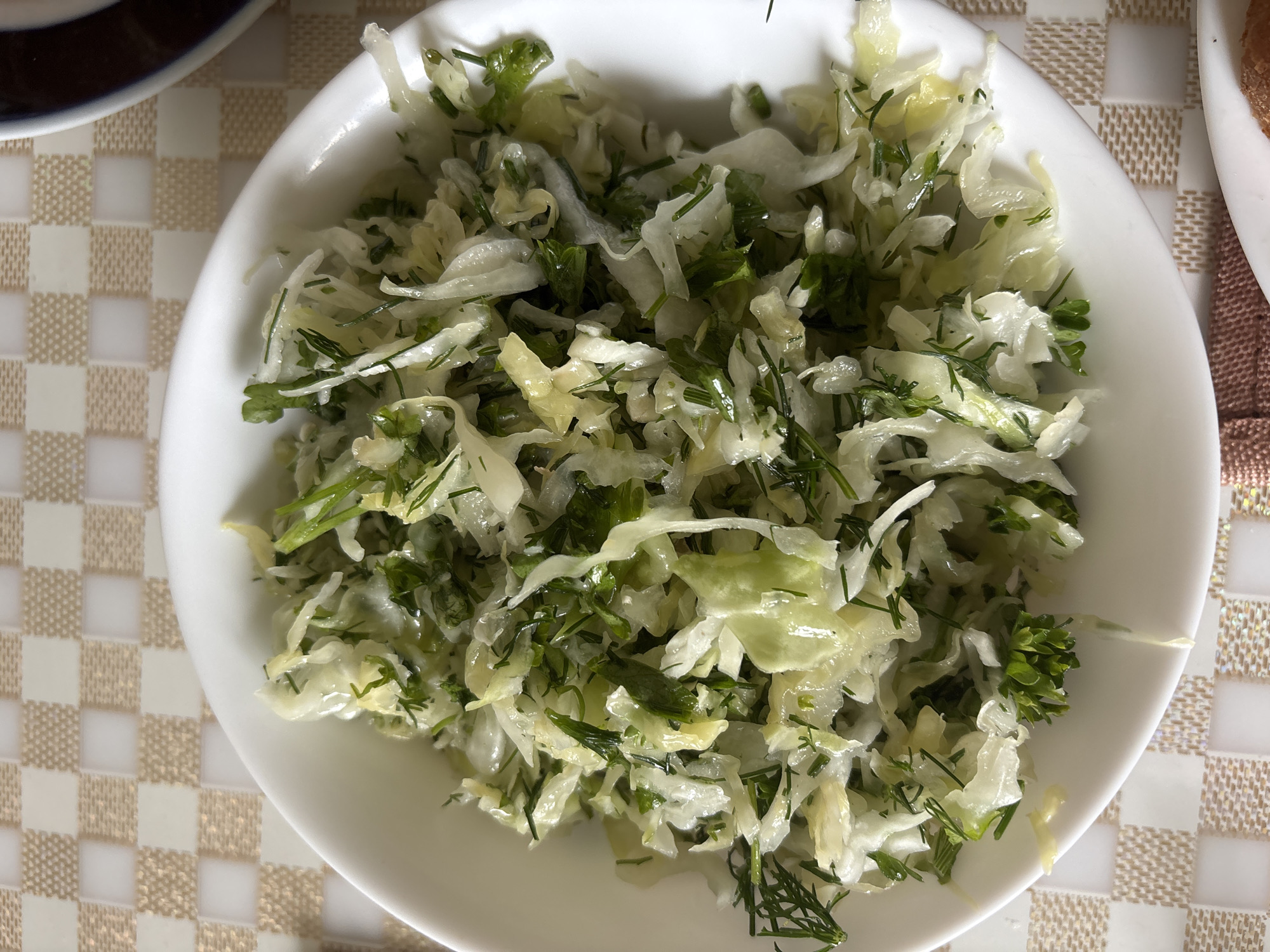 A white bowl of chopped cabbage salad with dill and parsley is placed on a checkered tablecloth. The salad appears to be lightly dressed. The lighting is natural and comes from above. The scene is static and no people are visible. The location is not specified in the image.