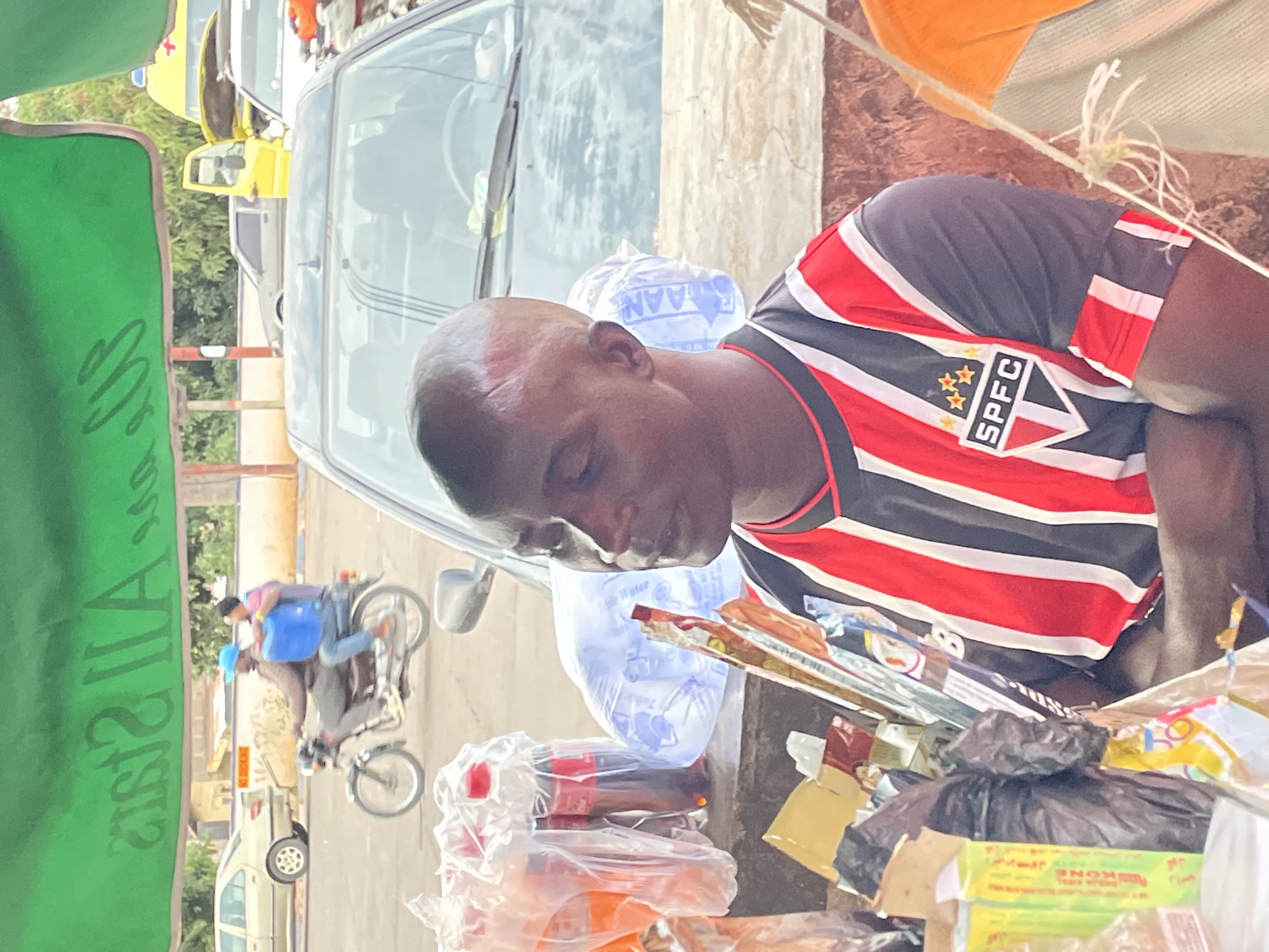 A man is standing next to a street-side display of goods. The man has a shaved head with a visible scar. He is wearing a soccer jersey with the letters "SPFC" and three stars on the chest. He is looking down at the goods on display. The goods appear to be packaged foods and other small items. In the background, there are cars, motorcycles, and other people on the street, with yellow taxis. The location is Jos, Nigeria.