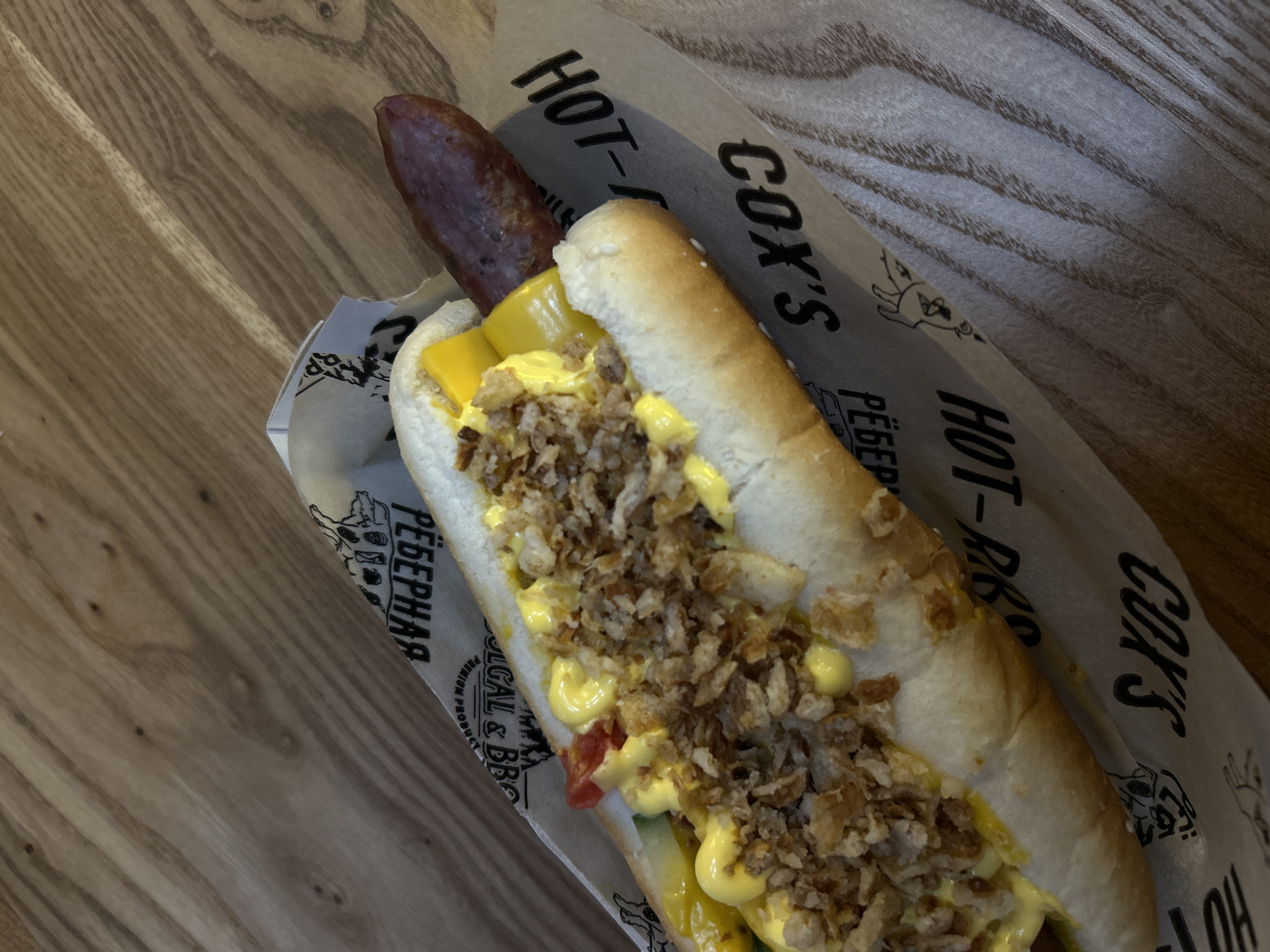 An overhead, eye-level, close-up shot shows a single, whole, uncooked beef frankfurter hot dog with various toppings on a wooden table. The hot dog sits on a piece of printed paper wrapping. The bun is split lengthwise, and the frankfurter is visible at one end, with the other end tucked inside. Toppings include dollops of yellow mustard, chopped red tomato, chopped green cucumber, and a generous amount of crispy fried onions. The paper wrapping has black text and images, including the words "HOT-R" and "COX'S" repeated. The background is a light brown wooden surface with visible grain patterns. The lighting is soft and even, creating minimal shadows.