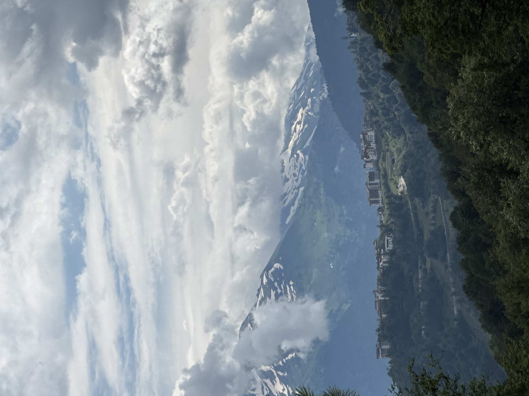 The image shows a mountain landscape with a resort village nestled on the slopes. In the foreground, dense green trees cover the lower parts of the mountains. In the midground, a cluster of white buildings, likely hotels or apartment complexes, are visible on a hillside. These buildings are spread out and appear to be part of a ski resort or mountain village. Further in the background, snow-capped mountains rise, partially obscured by dramatic, cloudy skies. The sky is filled with a mix of white and grey clouds, with some patches of blue visible. The overall scene suggests a view of a mountain resort in a location like Estosadok, Russia, known for its alpine scenery and winter sports facilities. There are no people or animals visible in the image.