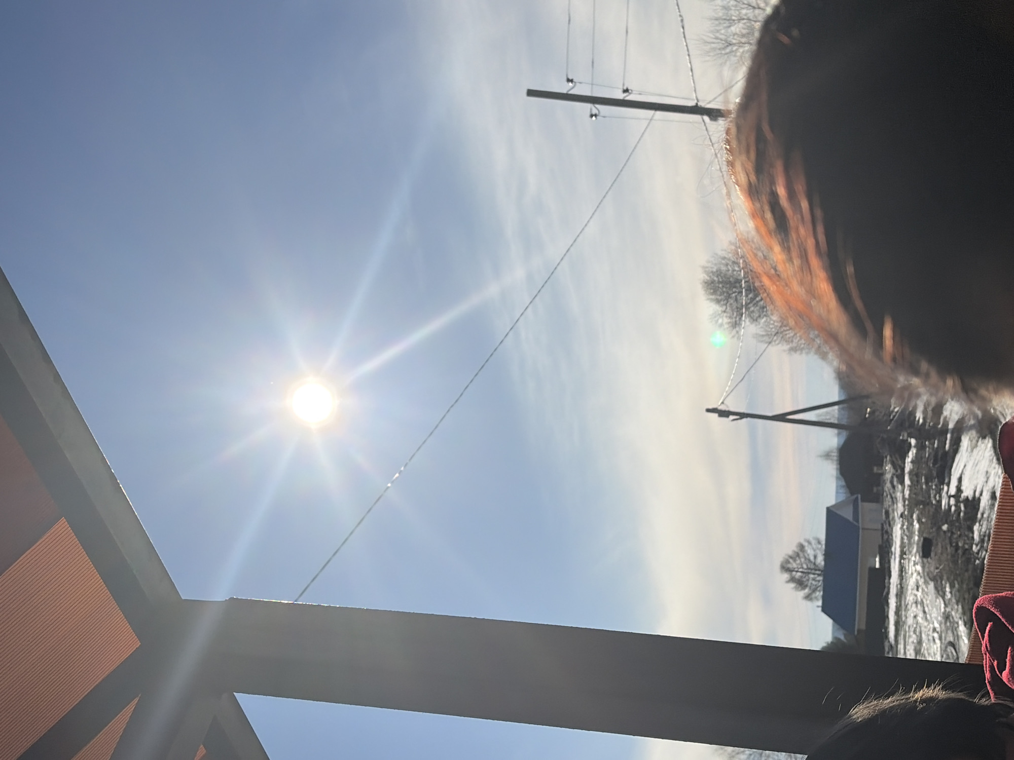 The image shows a bright, sunny sky with the sun prominently visible. A utility pole with power lines stretches across the upper portion of the image. The roof of a structure is visible in the bottom left corner. In the background, a house with a dark roof stands among snow-covered ground. A person's head is partially visible in the upper right and lower right corners. The location is consistent with a rural setting in Gory, Russia, given the presence of utility poles and residential buildings.