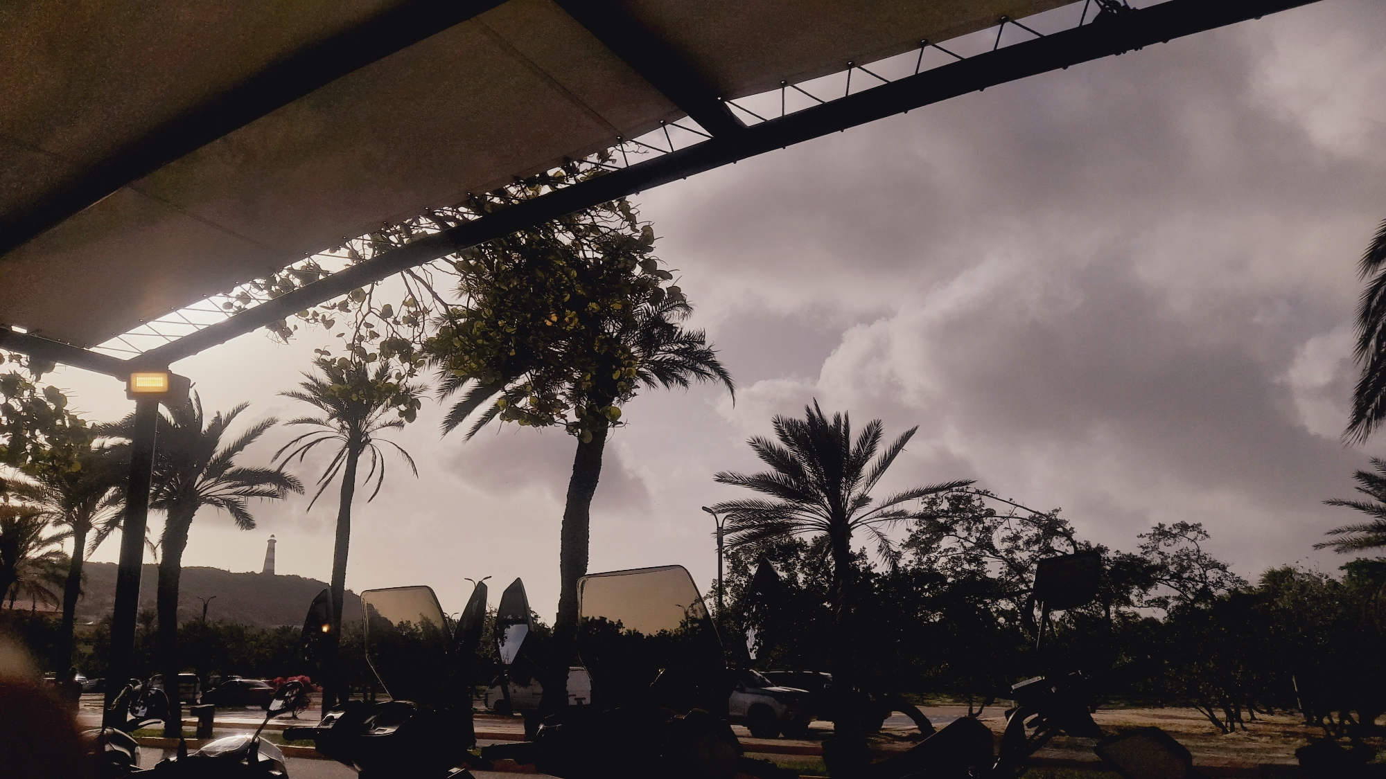 The image depicts an overcast sky over a coastal area, with silhouettes of palm trees and vegetation in the foreground. A lighthouse is visible on a hill in the distance. The scene is framed by a dark canopy structure in the upper portion of the image, with a yellow light fixture attached to its underside. Several motorcycles and their windshields are in the immediate foreground, suggesting an outdoor seating area or parking lot. The lighting indicates it is daytime, likely before or after a storm, given the heavy cloud cover. The overall impression is one of a tropical or subtropical location with a slightly muted and moody atmosphere. This scene is consistent with the environment found in Pedro González, Venezuela, a coastal town known for its lighthouse and palm-lined landscapes.