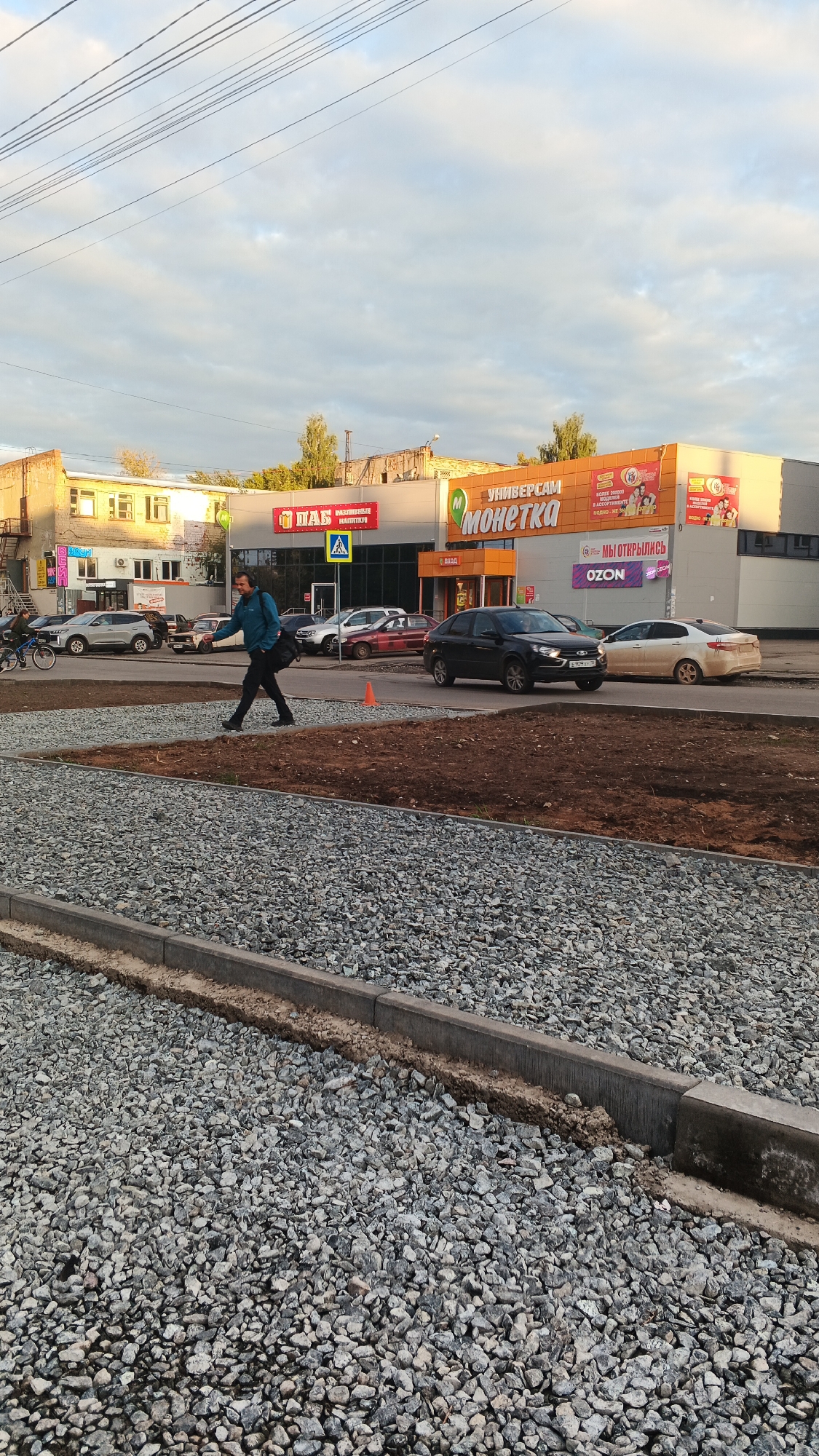 A man in a blue jacket and black pants walks across a gravel-covered area. He carries a black bag over his left shoulder and appears to be walking towards the right side of the frame. In the background, a commercial building with signs for "PAB" and "MONETKA" is visible. Several cars are parked and driving in front of the building. The sky above is cloudy. This scene is located on a street in Izhevsk, Russia.