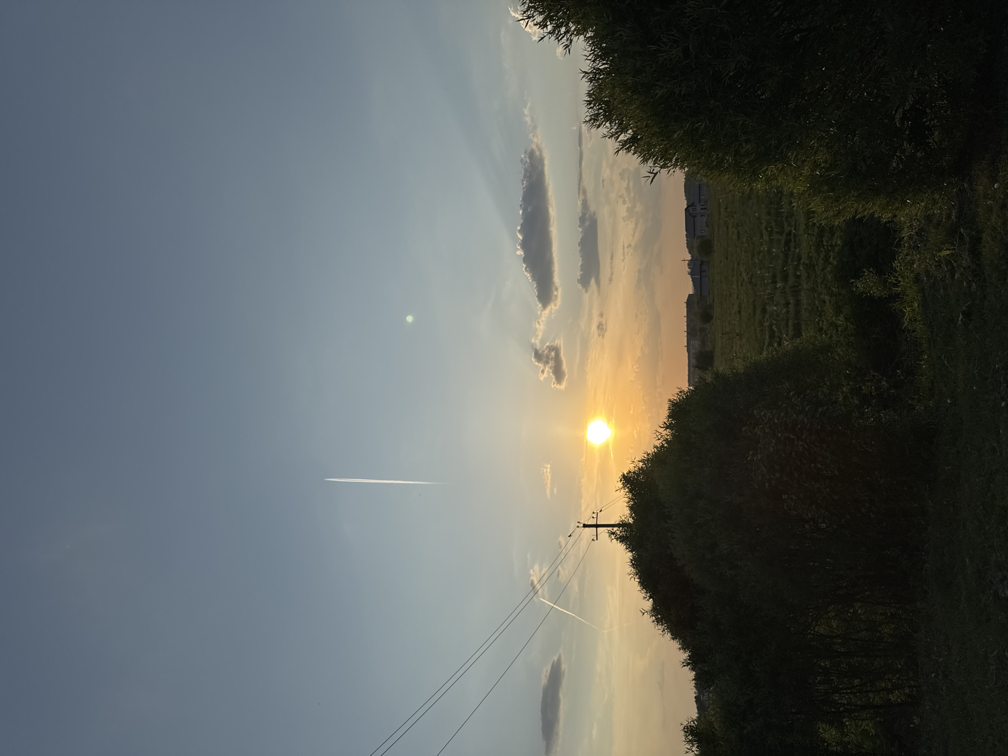 The image shows a landscape in Горный, Russia, at sunset. The sun is partially obscured by trees on the right side, creating a bright, golden glow in the sky. There are scattered clouds and contrails from airplanes in the blue sky. A utility pole and power lines are visible. In the distance, beyond the trees, there is a field and buildings.