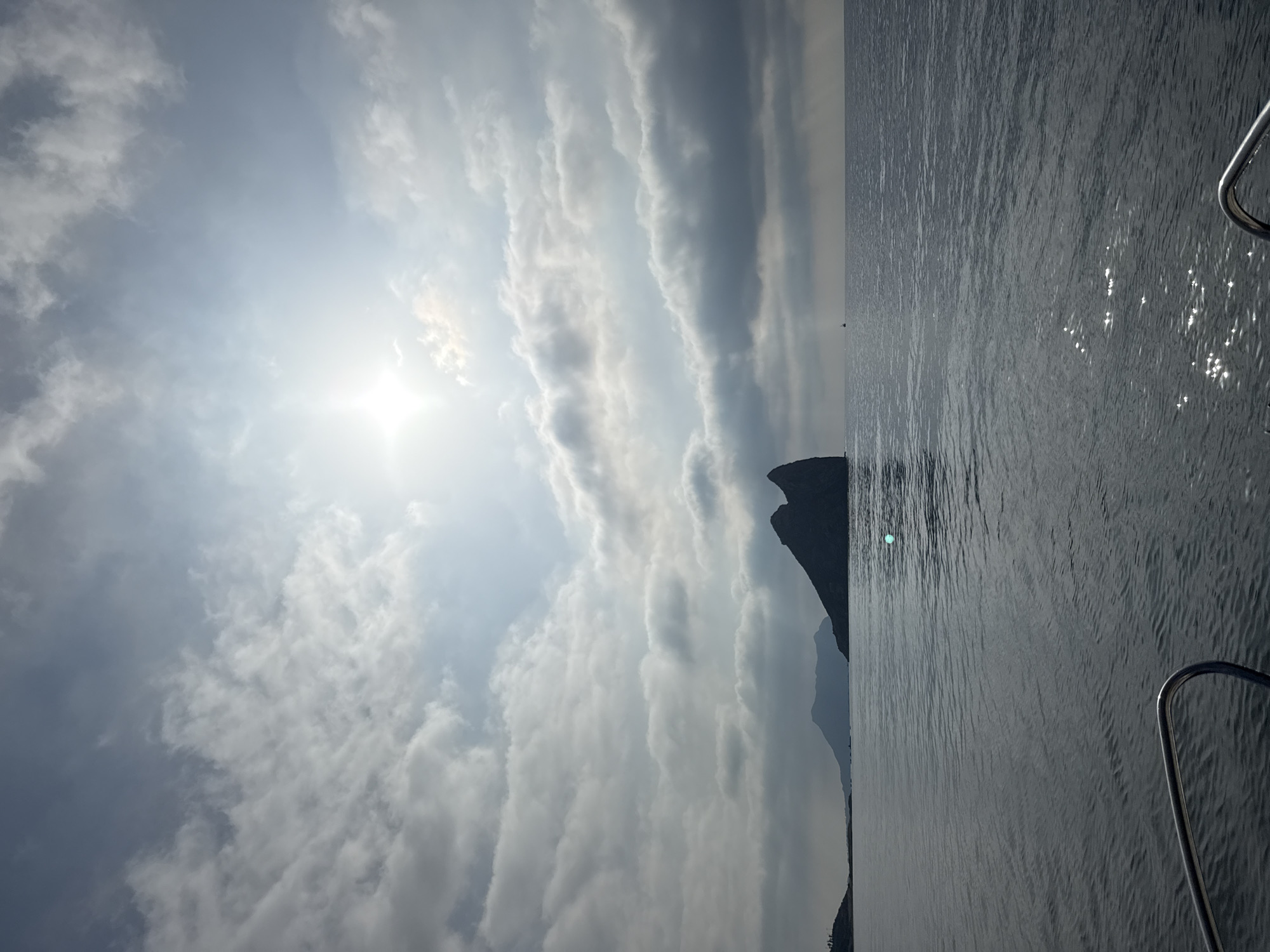 This is a photograph taken from a boat in the sea, looking towards the coastline of Águilas, Spain. The sun is visible in the upper right corner of the image, partially obscured by clouds. The sea is calm with light ripples. In the distance, silhouetted against the sky, are several rock formations and mountains. The closest rock formation is a prominent, dark, pointed mass that juts out of the water. Further inland, there are more mountainous landforms. The foreground of the image shows a metal railing from the boat. The overall scene is one of a seascape under a partly cloudy sky.