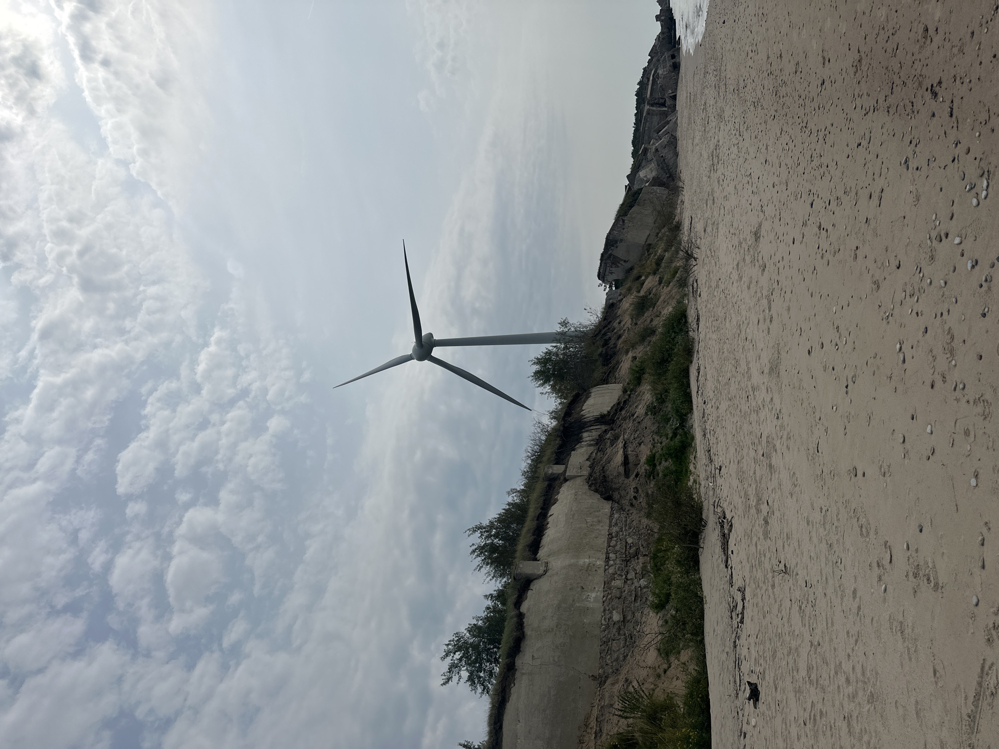 A wide-angle, eye-level shot captures a beach in Liepaja, Latvia, featuring a lone wind turbine standing tall against a cloudy sky. The foreground is dominated by a sandy beach with scattered pebbles and driftwood. To the left, a steep, eroded embankment is visible, covered with vegetation and a crumbling concrete structure. The wind turbine is positioned on top of this embankment, its three blades prominently displayed. In the background, the sea meets the shore, with more of the eroded embankment and concrete structures stretching along the coastline. The overall scene suggests a somewhat desolate yet picturesque coastal landscape, likely a remnant of wartime fortifications against the backdrop of renewable energy infrastructure. There are no people or animals present in the image.