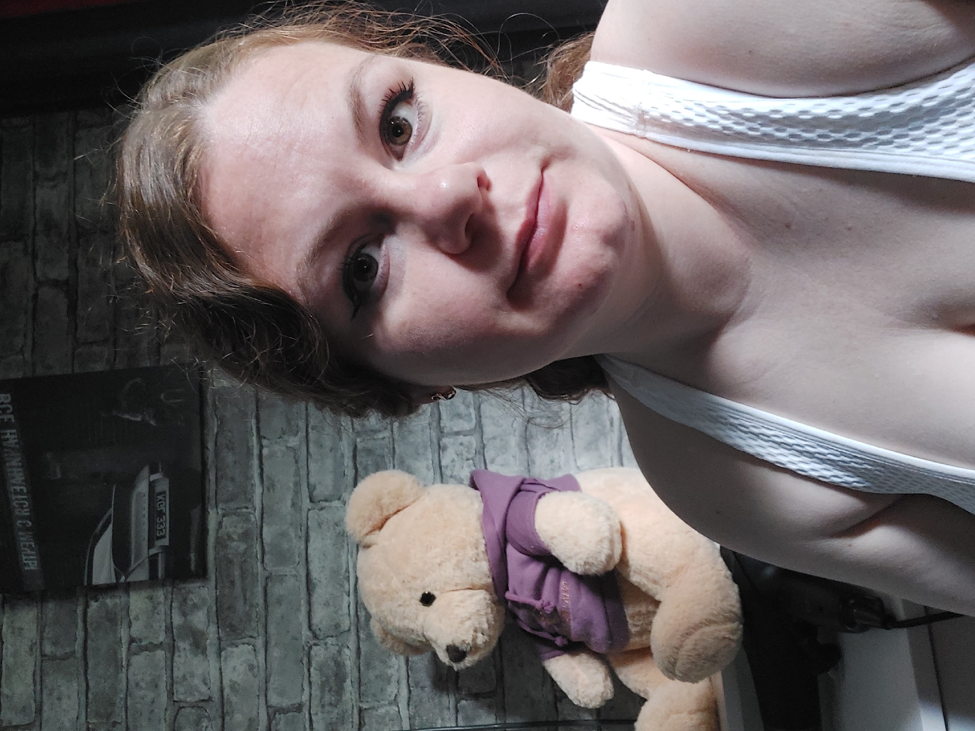 A selfie is taken from a close-up perspective, focusing on the face of a Caucasian woman with brown hair. She is wearing a white tank top. To her left, a large, light brown teddy bear wearing a purple hoodie is visible, sitting on a surface. In the background, a brick wall is present, with a framed picture of a car hanging on it. The lighting is dim, with a slight shadow cast on the right side of the woman's face.