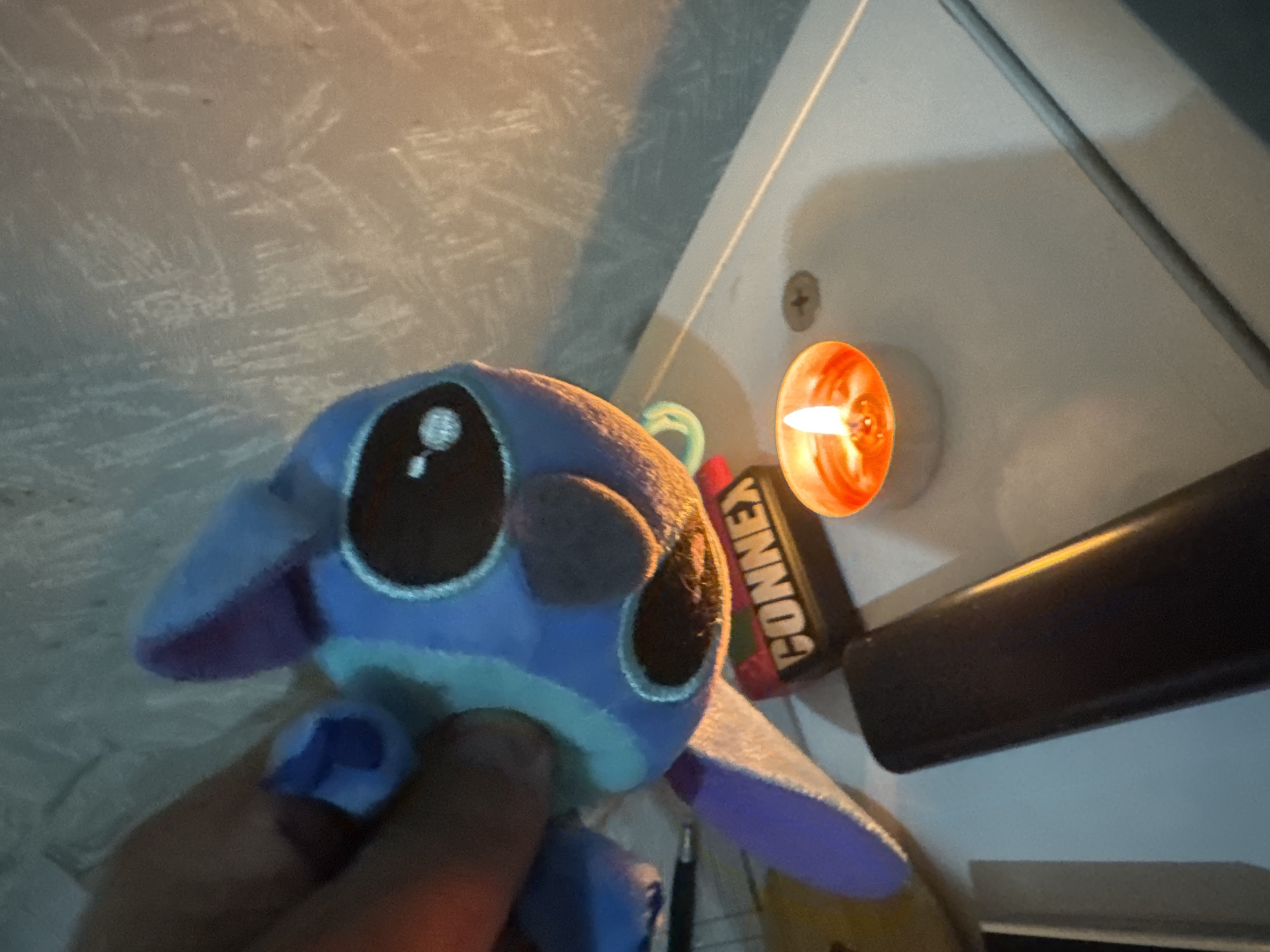 An upward-angled close-up shot captures a blue and purple plush toy, resembling Stitch from Disney's Lilo & Stitch, held by a person's hand in the foreground. The plush toy has large, dark eyes with white reflections, and a lighter blue stripe across its chest.
To the right and slightly behind the toy, a small, lit candle with an orange flame provides the primary illumination for the scene, casting a warm glow. Next to the candle, a dark-colored object, possibly a battery pack or tool, is visible, bearing the white text "CONNEX" on a red background. Further to the right, a black cylindrical object, resembling a marker or a small flashlight, rests on a light-colored surface. A metallic screw head is also visible near the candle on this surface.
The background features a textured, light-grey wall or surface with irregular patterns, suggestive of a stucco finish or a uniquely painted wall. Shadows are cast by the objects, indicating a single light source from the candle. The overall lighting suggests a dim environment, possibly evening or night, creating a soft and intimate atmosphere.
The scene appears to be an indoor setting, likely on a desk or shelf. The location context for this media is Sofia, Bulgaria.