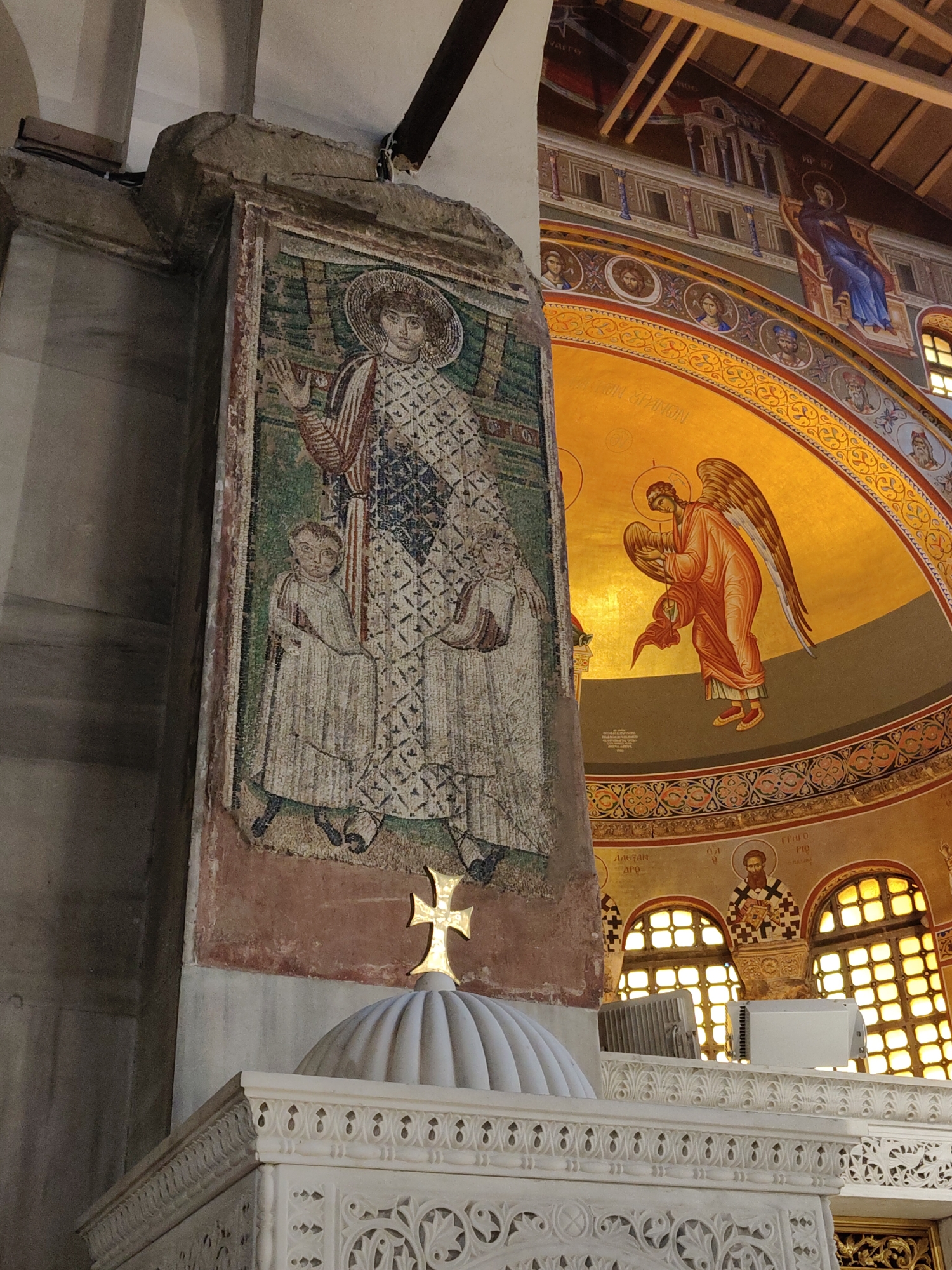 The image shows the interior of a Byzantine church in Thessaloniki, Greece. On the left, a large mosaic depicts a male figure with a halo and two children in white robes. The mosaic is affixed to a stone pillar. To the right, a gilded dome features a fresco of an angel with wings, dressed in an orange robe. Below the dome, a white marble structure with intricate carvings is visible, topped with a golden cross. In the background, arched windows with yellow glass illuminate the space, and further frescoes adorn the walls, including depictions of religious figures and architectural elements.