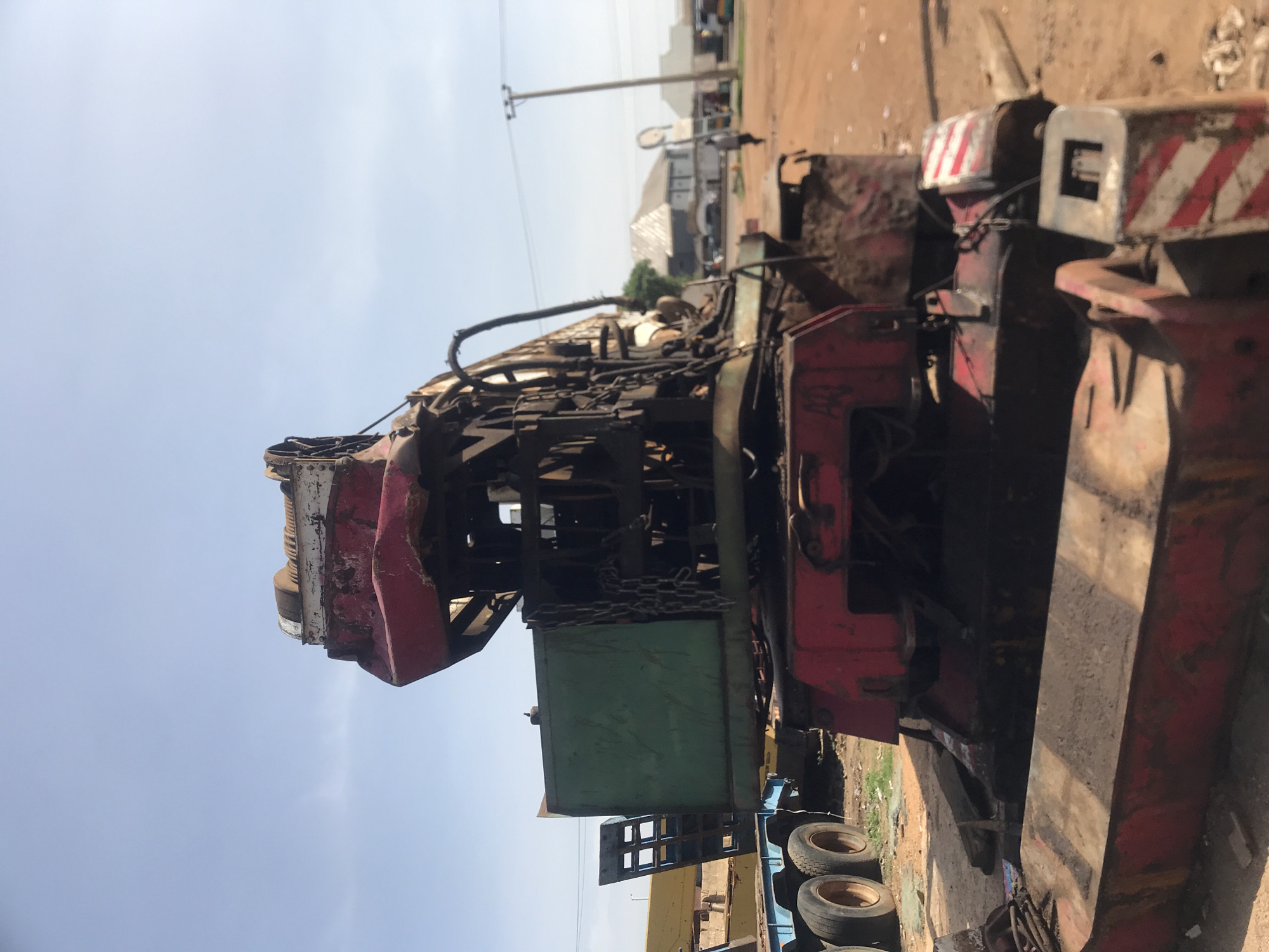 The image depicts a large, red and green crane, appearing weathered and possibly derelict, situated in an outdoor setting with a dirt ground. The crane has heavy machinery, chains, and a winch visible. In the background, there are buildings, a utility pole with wires, and a few indistinct figures. One figure, walking away from the camera, is dressed in dark clothing. Another vehicle, possibly a truck with blue and black tires, is partially visible to the left of the crane. The sky is clear and light blue. The scene suggests a possibly industrial or storage area within a town or city. The specific location in Rafin Par, Nigeria, indicates an urban or semi-urban environment. The overall impression is one of a functional or formerly functional piece of heavy equipment in a potentially active, though dusty, environment.