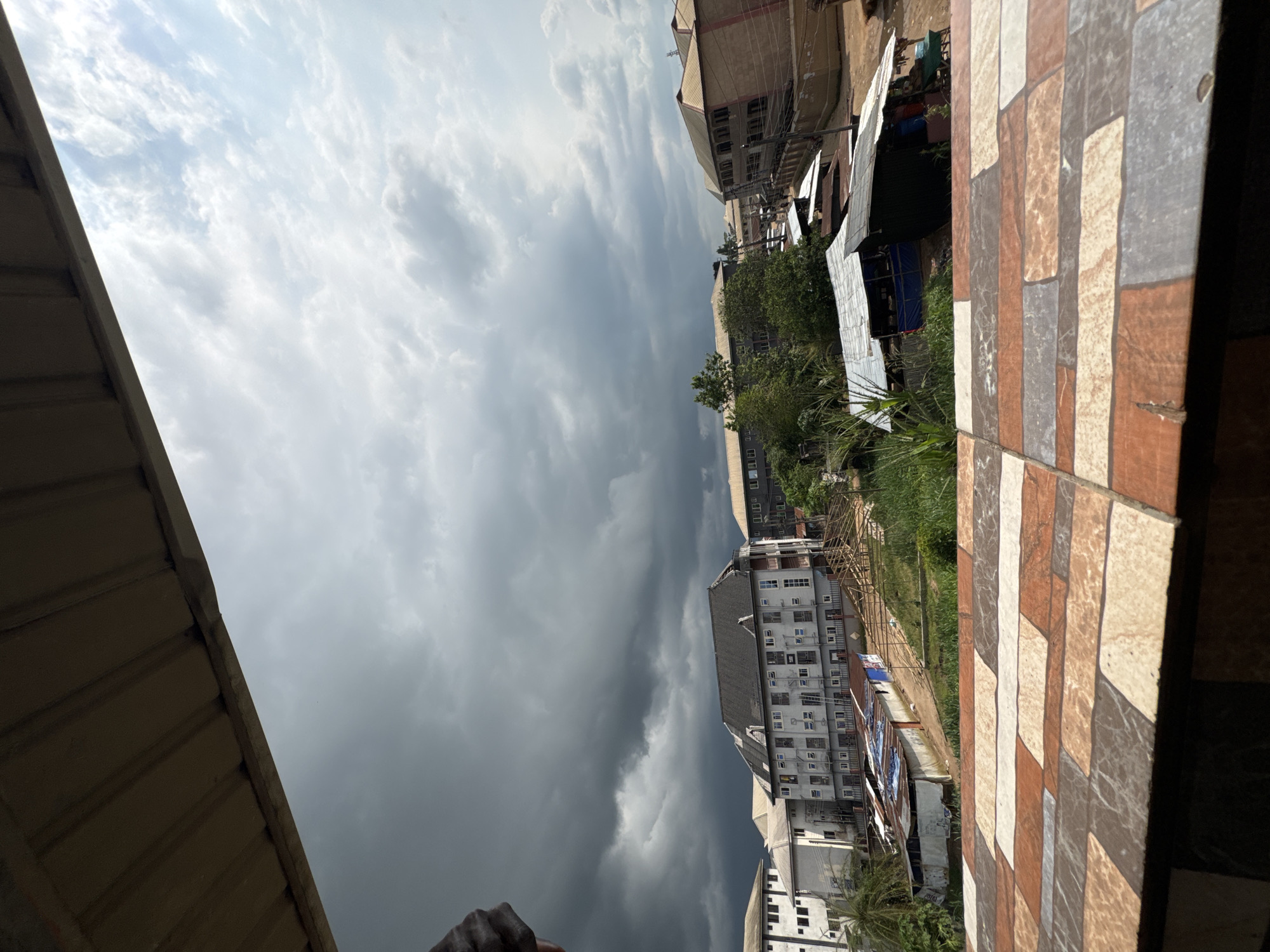 An overhead shot shows a view of Ihiagwa, Nigeria, looking out over a cluster of buildings and a sky filled with ominous storm clouds. The foreground is dominated by a tiled surface, likely a balcony or rooftop, with a patterned arrangement of brown, white, and grey rectangular tiles. Beyond this, the scene unfolds with several buildings, including a prominent, multi-story structure with a dark, pitched roof and numerous windows. To the left and right, other buildings of varying heights and colors are visible. Some appear to be residential, while others might be commercial establishments or educational institutions. Several smaller, more rudimentary structures with corrugated metal roofs are scattered throughout the midground, suggesting a mix of development. Lush green vegetation, including trees and tall grasses, intersperses the buildings, indicating a tropical or sub-tropical environment. The sky above is dramatic, with dark, heavy clouds on the left transitioning to lighter, scattered clouds and patches of blue sky on the right, hinting at an approaching or passing storm. There are no visible people or animals in the image.