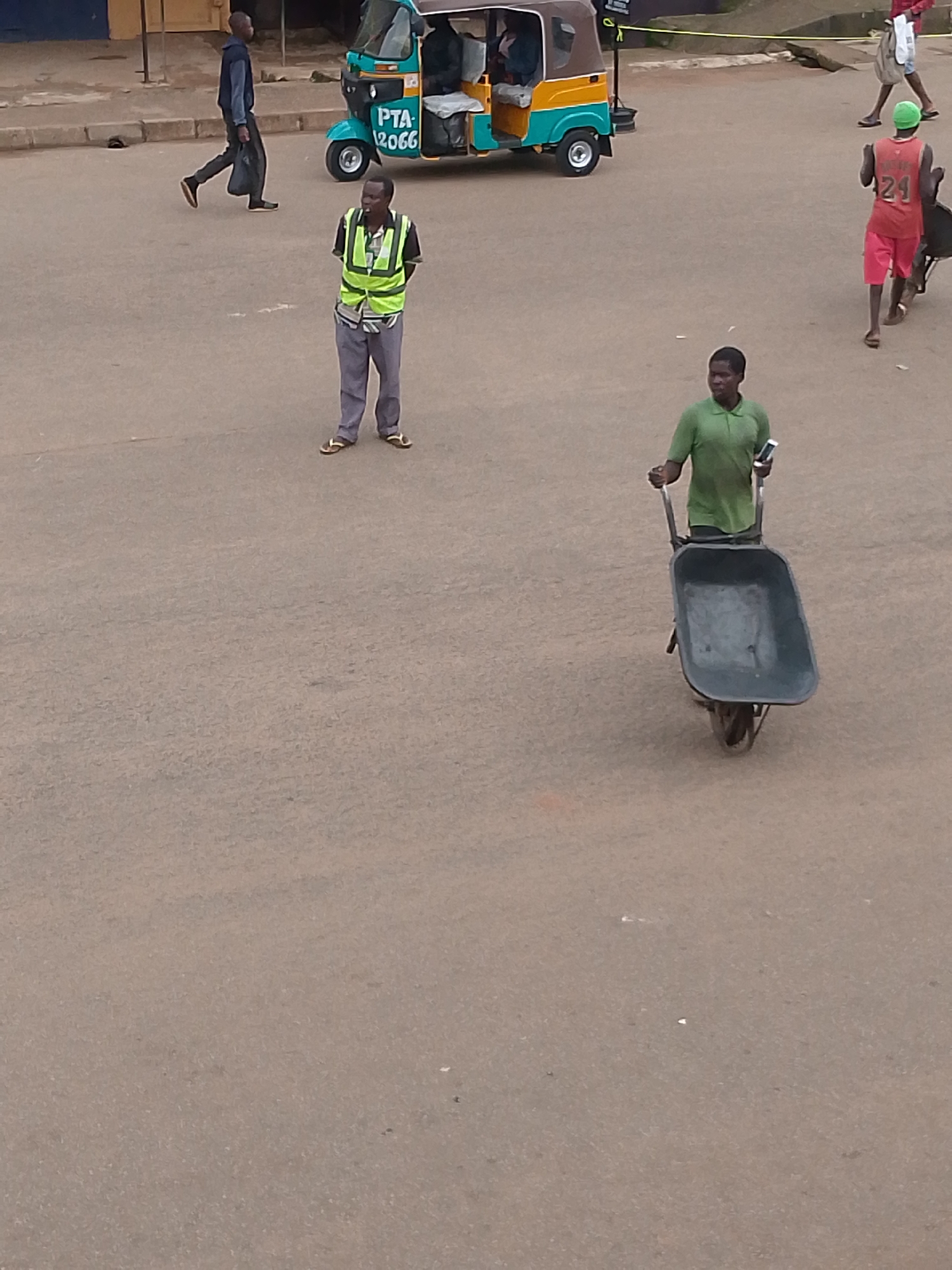 In Bukuru, Nigeria, two men are observed on a paved road. The man on the left is wearing a reflective green vest over a blue shirt and grey pants, with his hands behind his back. He is standing in front of a teal and yellow auto-rickshaw, which has "PTA 2066" written on its side. To the right, another man is pushing an empty, dark-colored wheelbarrow, holding a white cell phone in his right hand. He is wearing a green polo shirt and appears to be walking away from the viewer. In the background, a man in a red shirt and pink shorts with a green cap is visible. The road surface is a neutral gray asphalt, with some debris and smudges visible. The lighting suggests it is daytime.