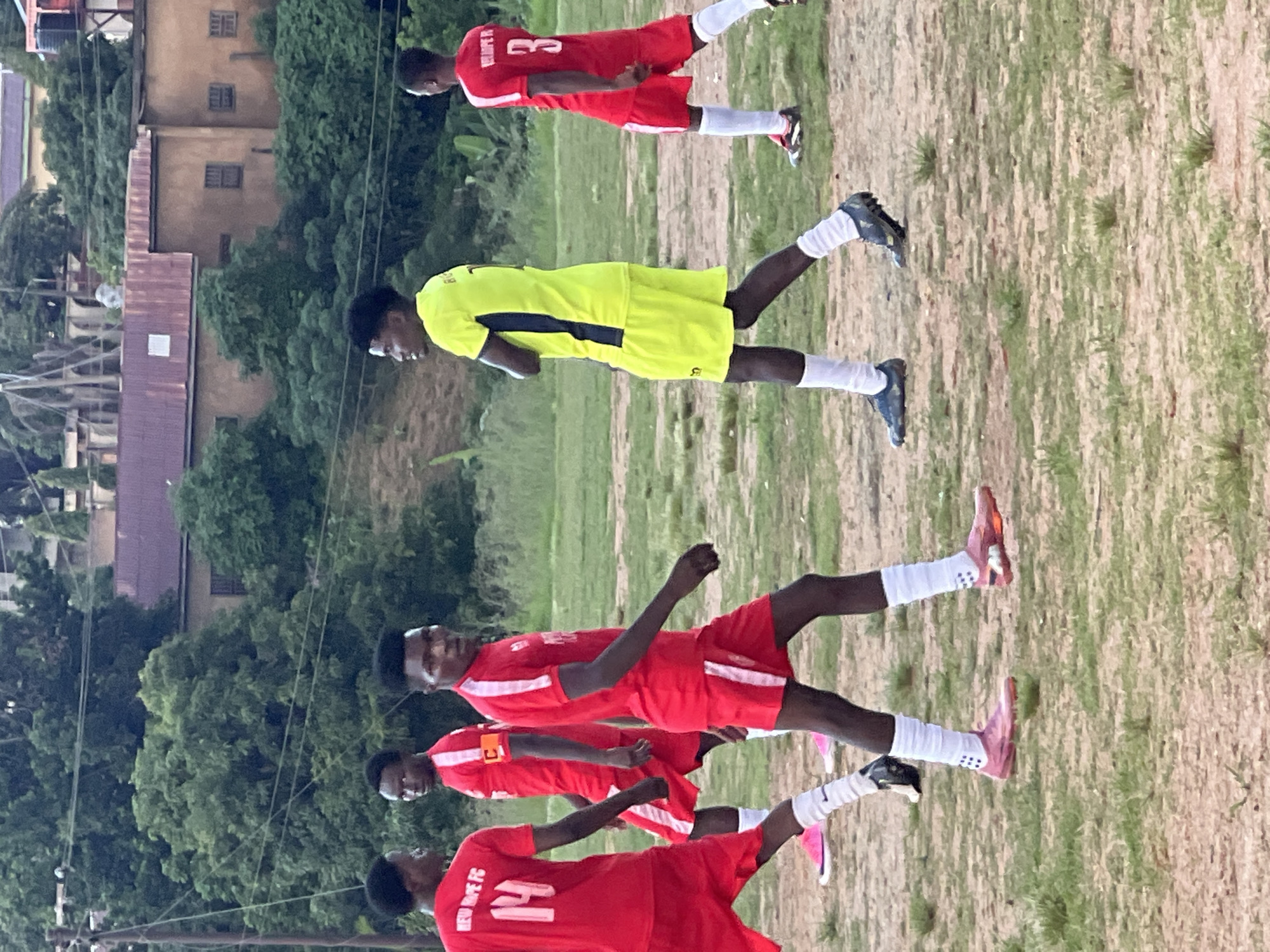 A group of male soccer players are on a dirt field in Jos, Nigeria. The players are wearing red and yellow jerseys with white shorts and socks. Some players have the number 14, 3, and C on their jerseys. They are facing in different directions, and some appear to be walking or jogging. The field is surrounded by trees and buildings. In the background, there are several buildings, some with corrugated metal roofs. There are also telephone poles and wires visible. The sky is overcast.