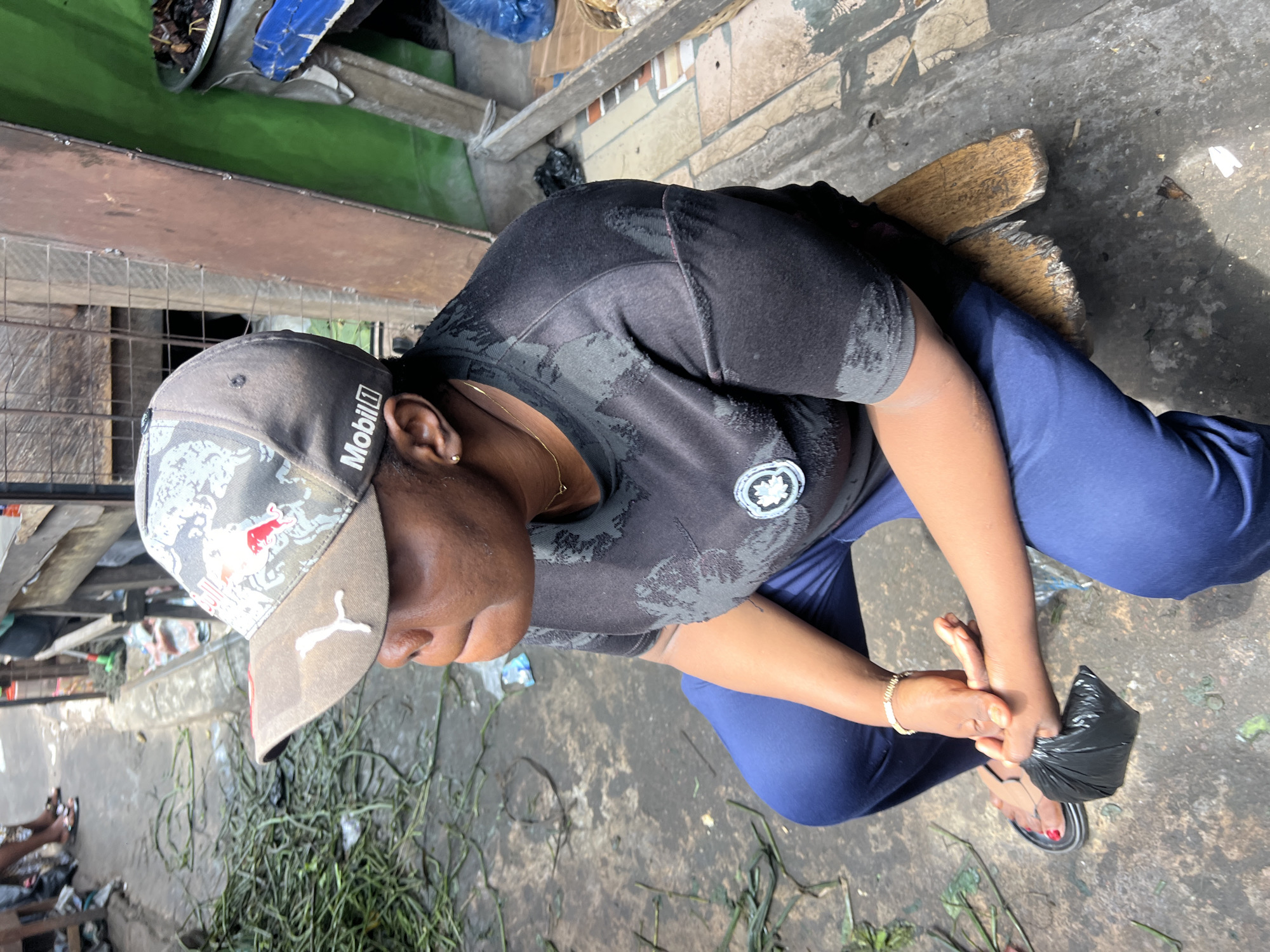 A medium shot shows a Black woman sitting on a wooden bench in Nkpor, Nigeria. The woman is wearing a black t-shirt and blue pants. She has on a grey baseball cap with a red and white logo on the front. Her arms are crossed and she is holding a small black bag in her hands. The background of the image shows a street with a dirt ground. There are some green plants on the ground and a wooden structure in the background.