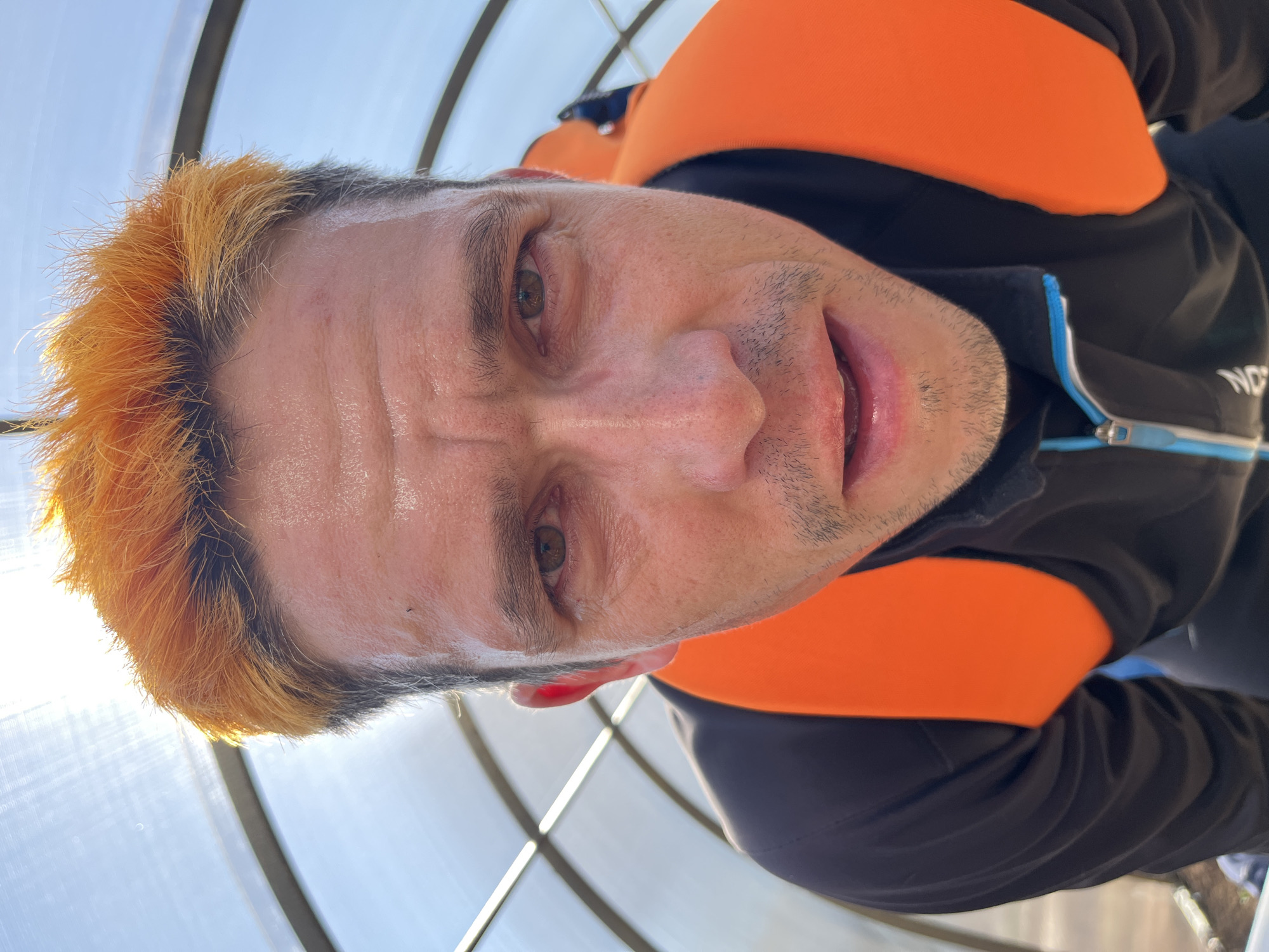 A close-up, angled portrait of a man with orange-tinted hair, captured in Chaikovsky, Russia. He is wearing a black and orange jacket. The background consists of a translucent, arched structure. The man appears to be sweaty or has a sheen on his face.