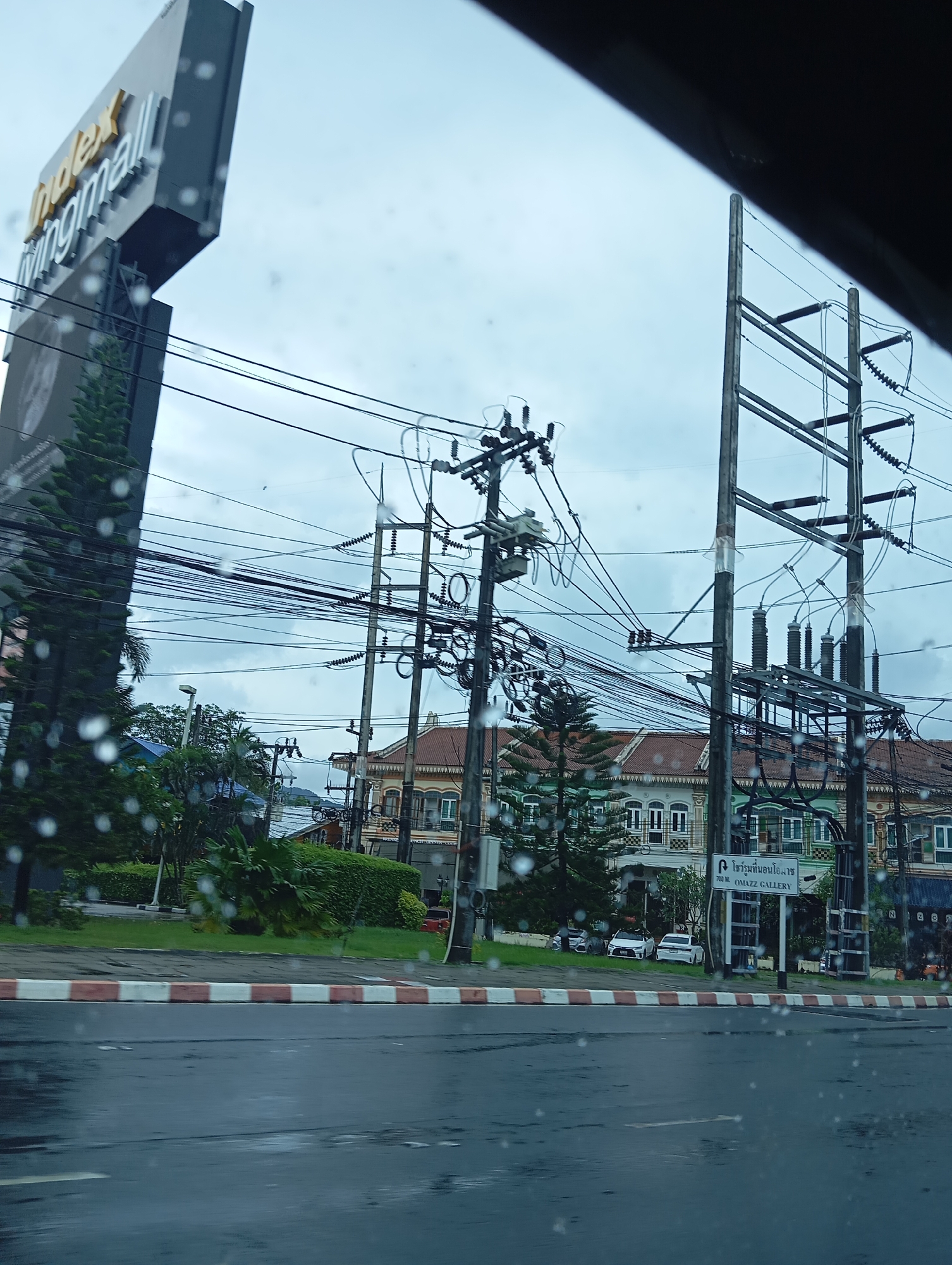 In a city street scene, likely in Thailand due to the signage, a large, tall sign with the words "Index Livingmall" is visible on the left. The sky is overcast, and there are raindrops on the camera lens, suggesting it is raining. In the foreground, a wet asphalt road stretches into the distance, bordered by a curb with red and white stripes. Behind the curb, a row of green hedges and trees lines the street. Across the street, a row of buildings with ornate architecture, featuring arched windows and tiled roofs, is visible. Electrical poles with a complex network of wires and transformers are prominent throughout the scene, indicating a busy urban environment. Some vehicles are parked along the roadside in front of the buildings. A signpost in front of one of the buildings indicates "700 M. OMAZZ GALLERY". The overall impression is of a rainy day in an urban area, with a mix of commercial and residential buildings.