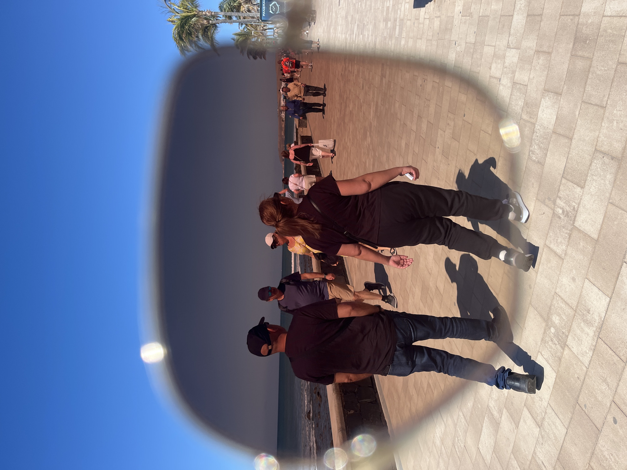 The image is taken through the lens of sunglasses, showing a partly sunny day at San Bartolomé de Tirajana, Spain. A section of the clear blue sky is visible to the left of the sunglasses frame. Within the lens's view, a paved walkway stretches along the coast. Several people are strolling along the walkway, including a man wearing a black shirt, blue jeans, and a baseball cap, and a woman with her hair in a ponytail, wearing a black shirt and black pants. There are other individuals further down the walkway, appearing as smaller figures due to distance. Palm trees are scattered along the walkway. In the background, there is a clock labeled "PANERAI". The ocean is visible between the walkway and the beach.