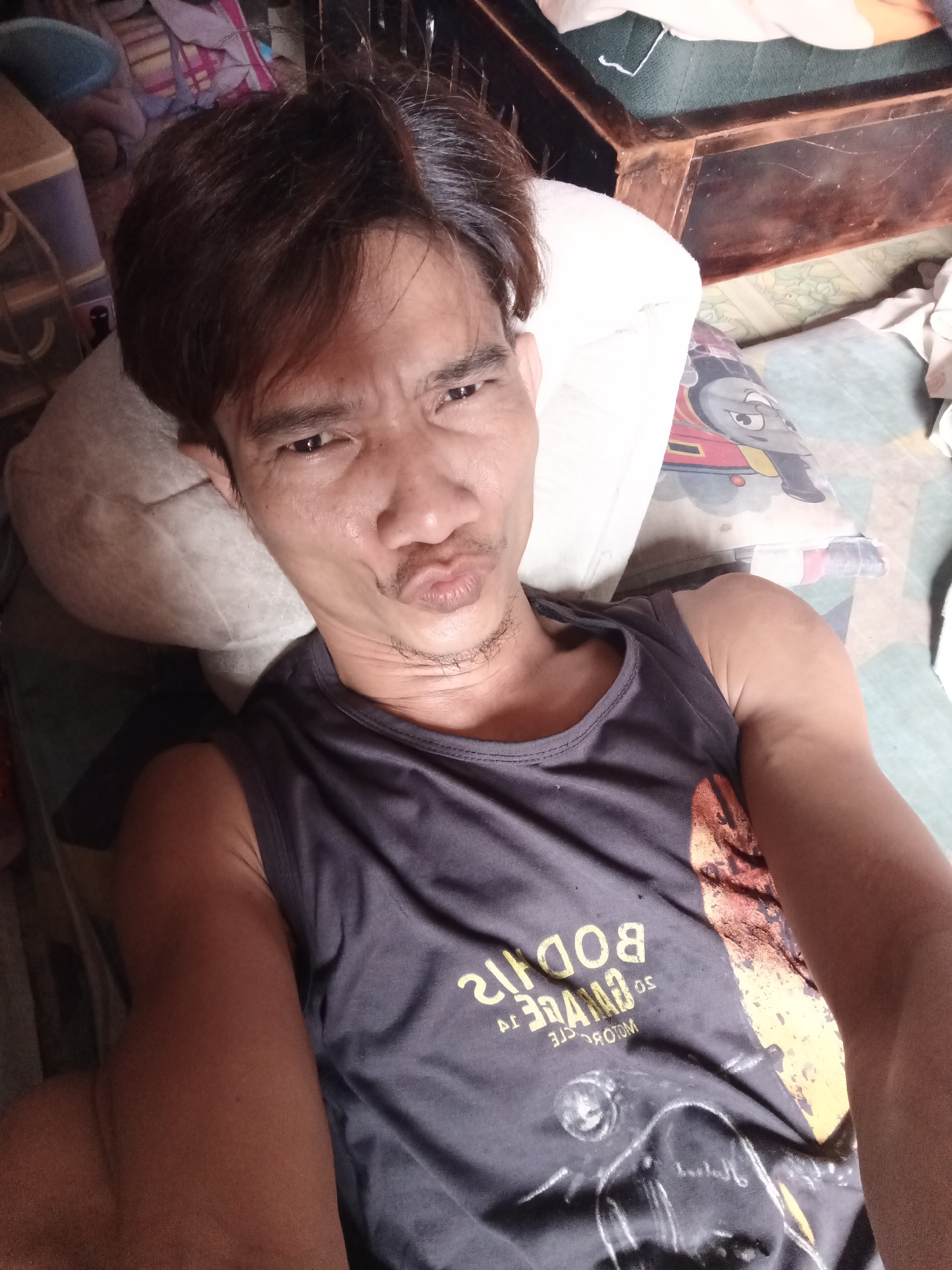 A man is lying down, looking at the camera with his mouth puckered. He is wearing a dark sleeveless shirt with yellow text and an image on it. The man has dark hair and stubble. He is lying on a bed with pillows and blankets. The background is out of focus and appears to be a bedroom. The scene is indoors. The location is not specified.