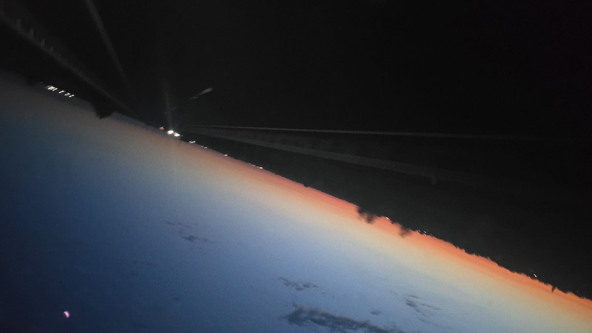 The image depicts a highway scene at dusk, with a clear view of the horizon transitioning from a deep blue sky to a gradient of orange and pink. The road appears to be a multi-lane highway, with white lane markings visible. There are faint lights of distant vehicles and structures along the side of the road. A small, crescent-shaped object, possibly the moon, is visible in the upper right corner of the sky, tinged with pink. The overall impression is one of a peaceful journey at the end of the day, with the urban landscape of Kövegy, Hungary, dimly visible in the distance.