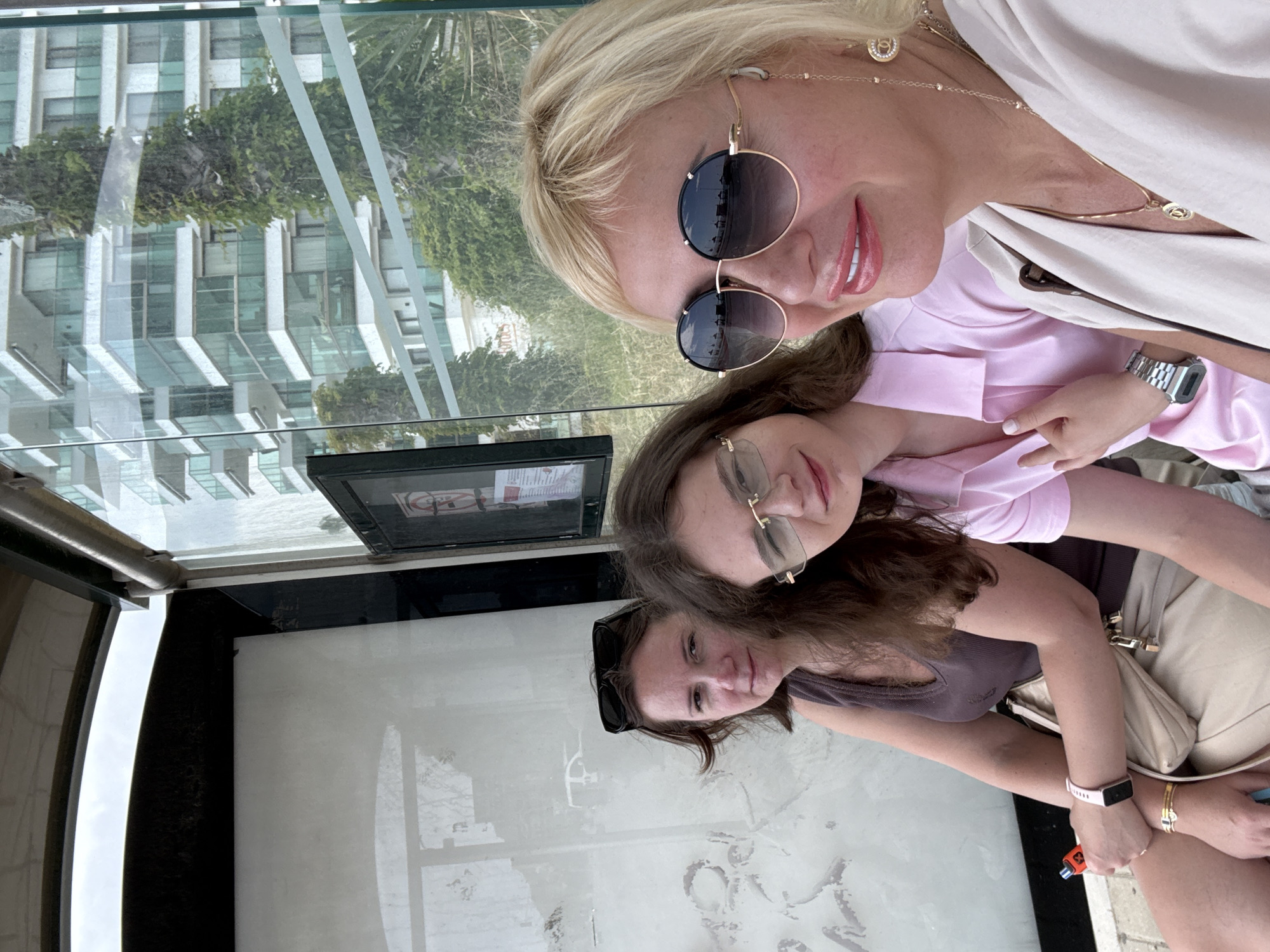 Three women are in what appears to be a bus shelter. A blonde woman with sunglasses is in the upper right of the frame. Below her is a woman with glasses and brown hair. In the lower left is a brunette woman with sunglasses perched on her head. The women are embracing and facing the camera. Through the glass of the shelter, a building and trees are visible.