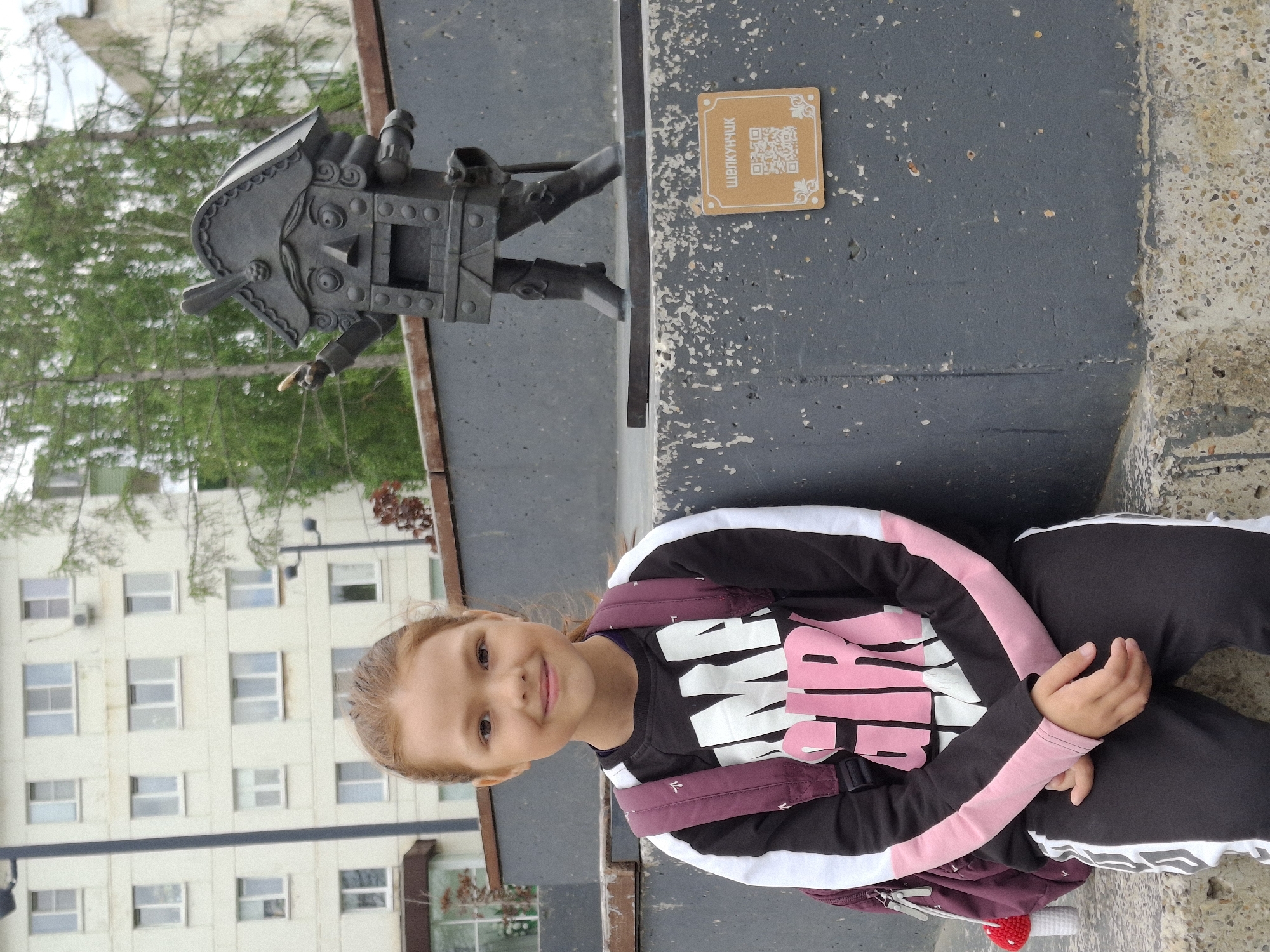 A young girl with auburn hair and fair skin sits on concrete steps in front of a statue of a nutcracker. She is wearing a black and white sweatshirt with pink sleeves and black sweatpants with white stripes. A maroon backpack with a small crocheted mushroom charm hangs on her left side. The nutcracker statue, made of dark metal, stands on a pedestal behind her. A small, square plaque with a QR code and Russian text, "ЩЕЛКУНЧИК" (Shchelkunchik, meaning Nutcracker), is affixed to the pedestal. In the background, a multi-story building with numerous windows is visible, along with trees. The scene appears to be outdoors, likely in a public space within the city of Chaikovsky, Russia, given the context of the nutcracker statue and its associated signage.