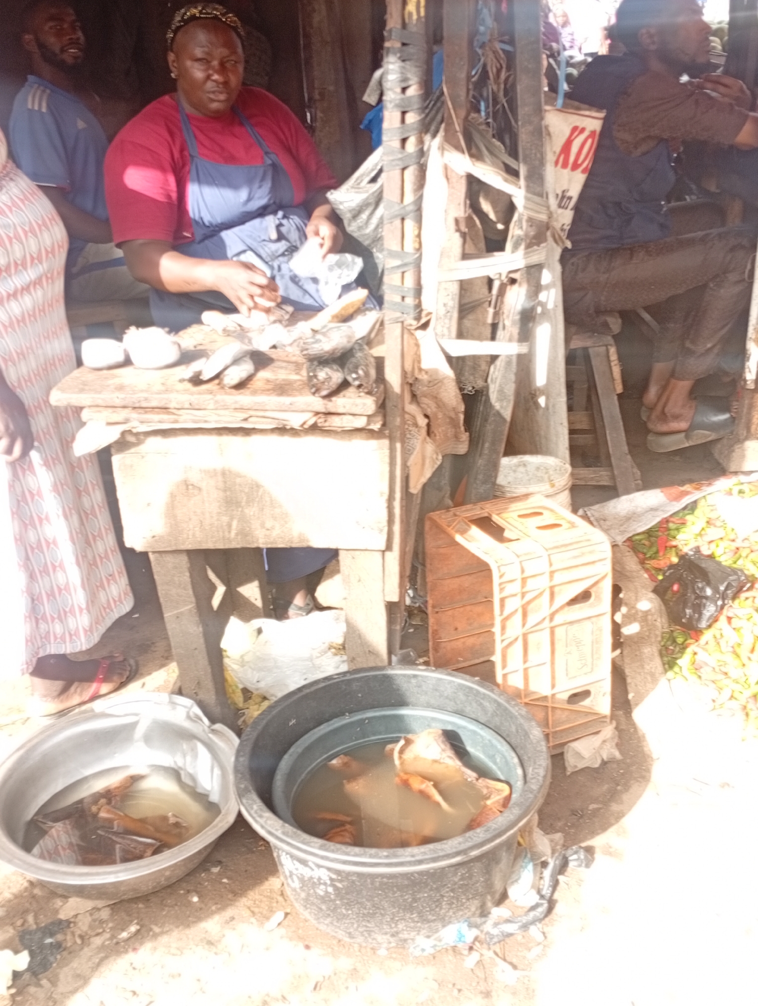 The image depicts a street market scene in Rafin Par, Nigeria. A woman wearing a red shirt and blue apron is preparing fish on a wooden table. Several fish are laid out on the table, with some appearing cleaned and others whole. To the left, a person in a patterned dress stands by. In the background, a man in a blue t-shirt is visible. Further to the right, another man, wearing a dark shirt and trousers, is seated. Various objects are scattered around the market, including plastic crates, buckets, and produce. The environment appears to be outdoors, with natural lighting and a rough, unpaved ground surface.