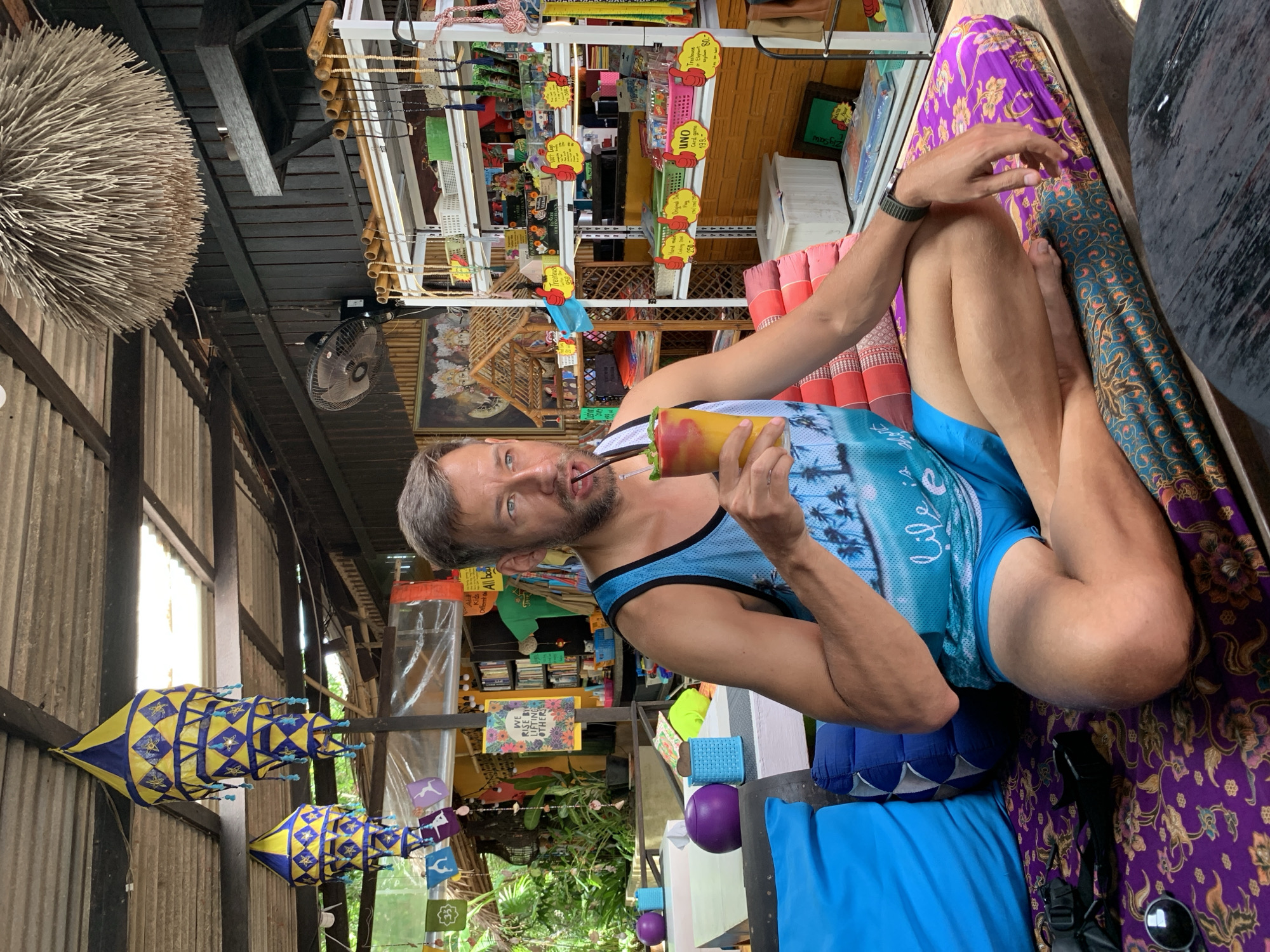A man with short brown hair and a light beard sits cross-legged on a cushioned bench in an outdoor, open-air establishment, likely a cafe or shop, in Baan Bo Phut, Thailand. He wears a blue tank top with a palm tree and ocean scene and blue shorts. The man is drinking from a tall glass filled with a layered orange and yellow beverage, with a straw inserted. The bench he sits on is covered with a patterned purple and blue fabric. Behind him, shelves are stocked with various items, including what appear to be handmade crafts and souvenirs. Decorations such as paper lanterns and a large, spherical hanging made of twigs are visible on the ceiling. The background suggests a tropical environment, with lush greenery visible. The overall atmosphere is casual and relaxed.
