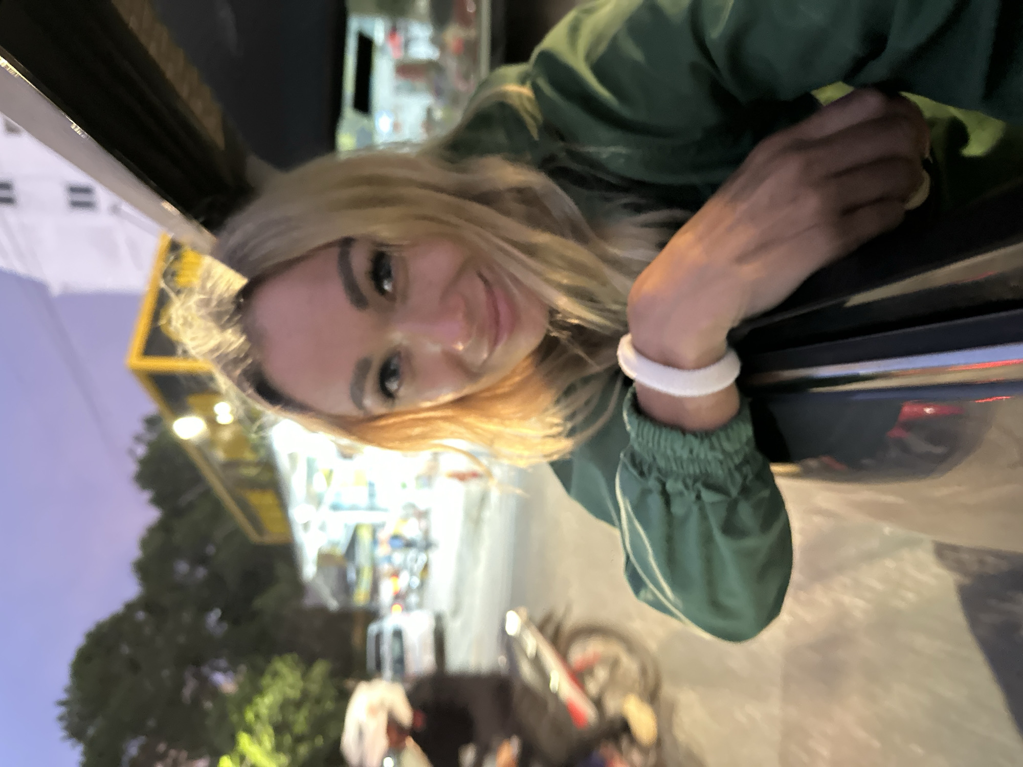 The media file shows a young woman with blonde hair, wearing a green jacket, leaning out of a vehicle window. She is smiling at the camera, and her right arm is resting on the car door. She has a white bracelet on her right wrist. The background is blurred, but appears to be an outdoor setting at dusk or night, with lights from buildings and possibly streetlights visible. There are trees and what looks like a yellow bus or structure in the distance. The overall mood is casual and happy. The specific location is Da Lat, Vietnam.